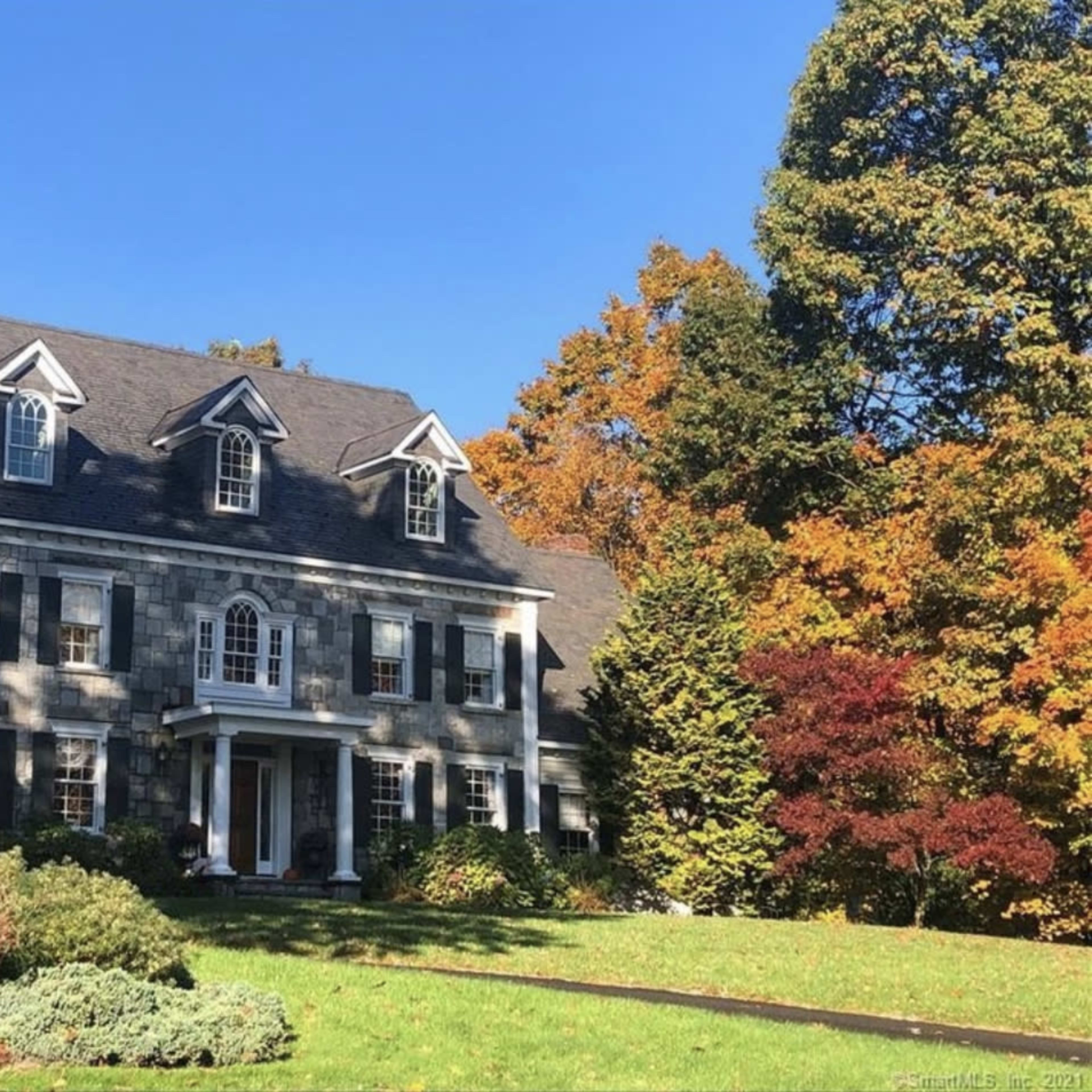 A large, two-story gray house with a symmetrical facade, surrounded by colorful autumn trees and well-maintained landscaping.