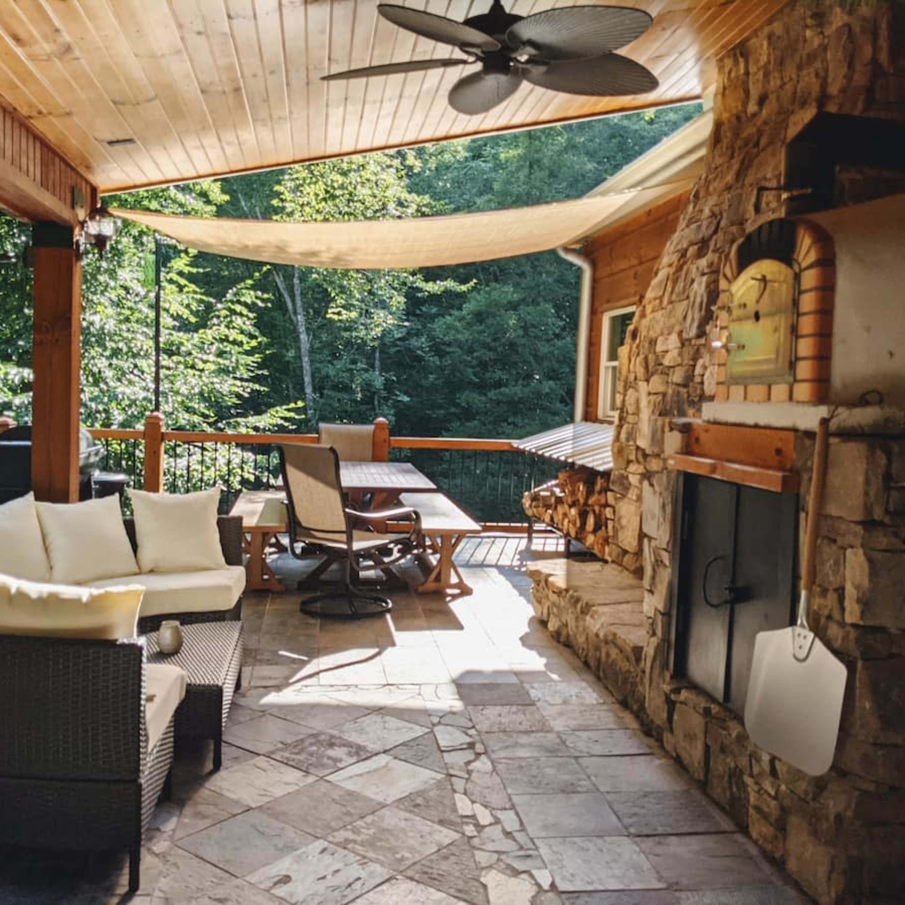 A covered outdoor patio featuring a stone fireplace, a dining table, and a cushioned seating arrangement, surrounded by greenery.