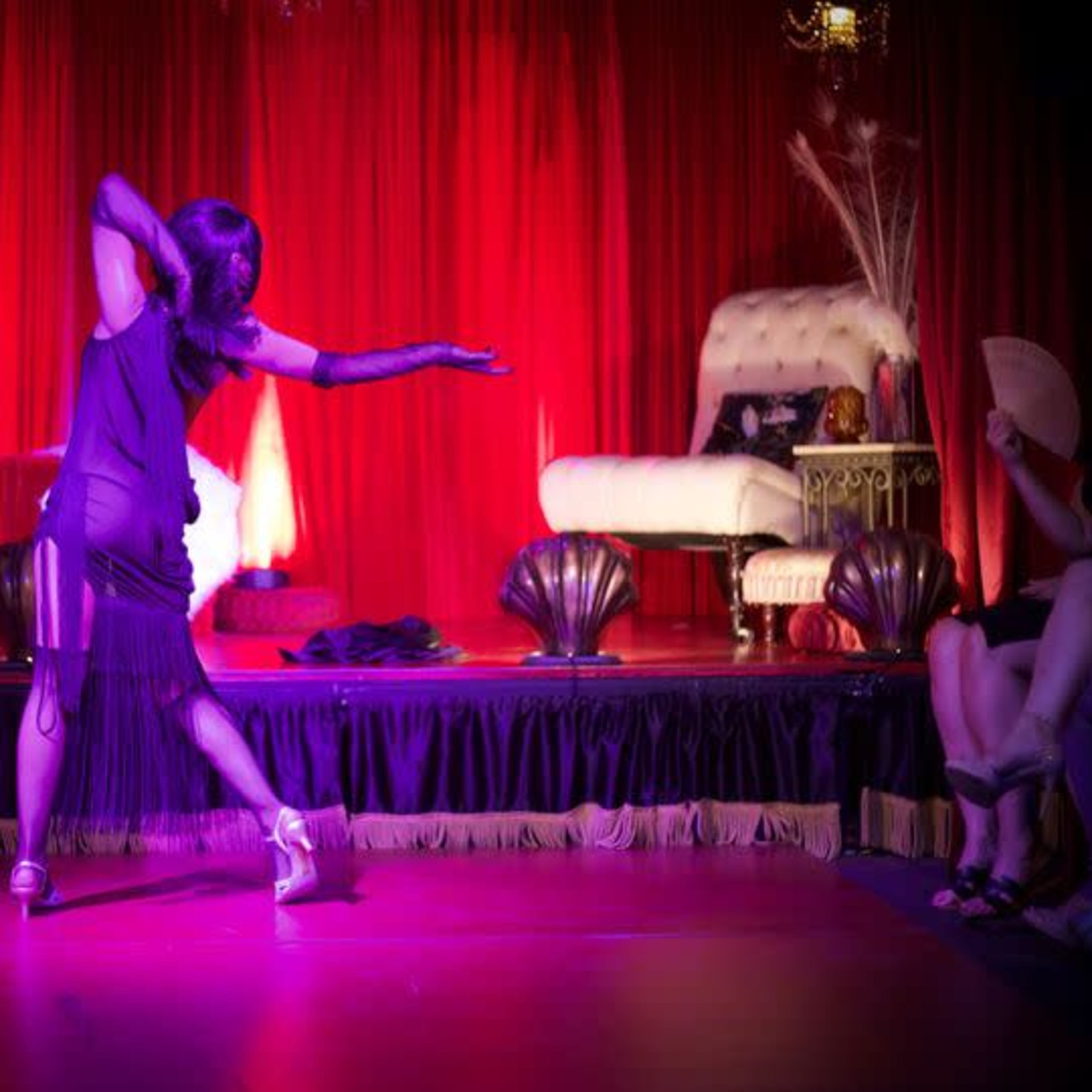 A dancer performs on stage in front of a red curtain, with a chaise lounge and audience seated nearby.
