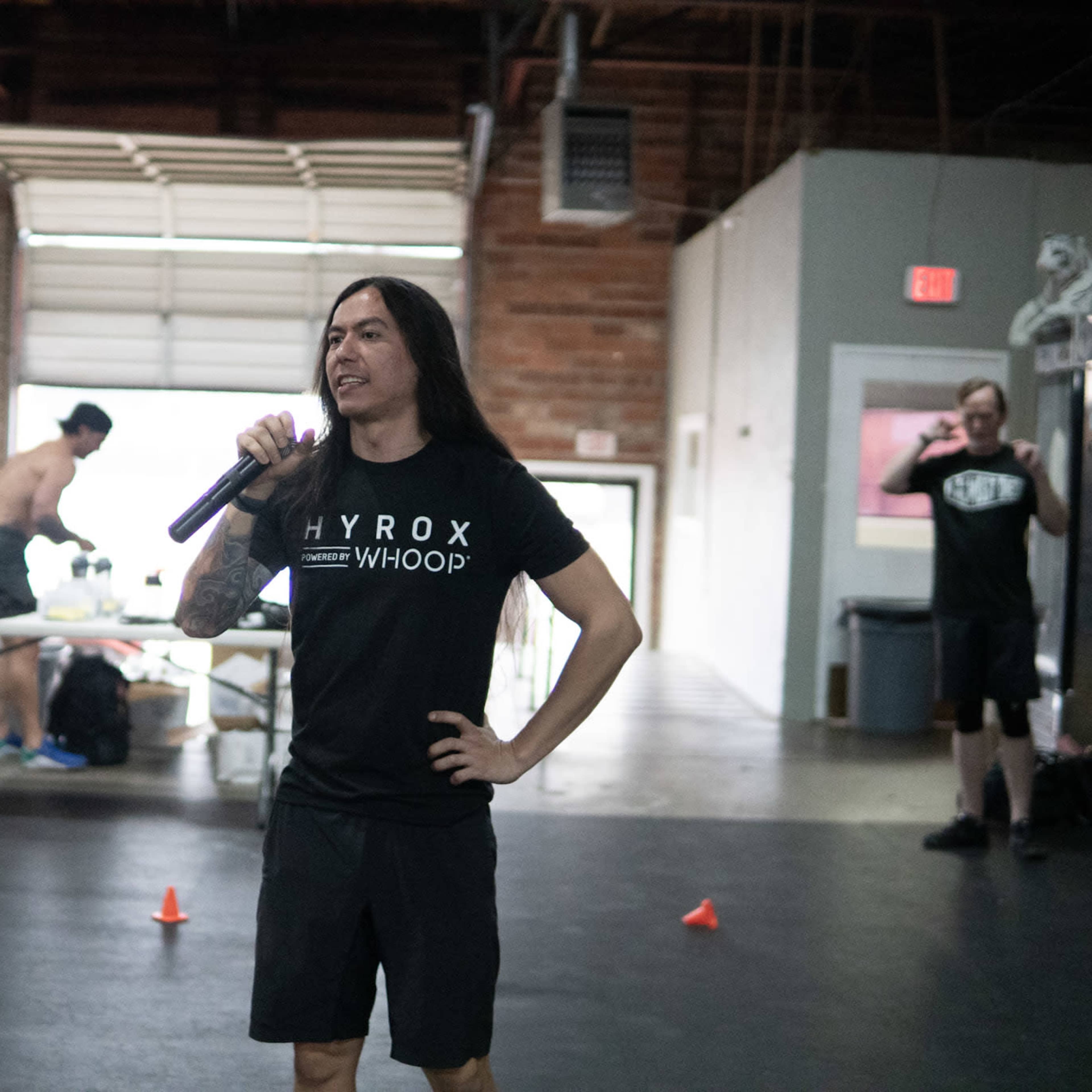 A person in a black t-shirt stands in a gym, holding a microphone while others train in the background.