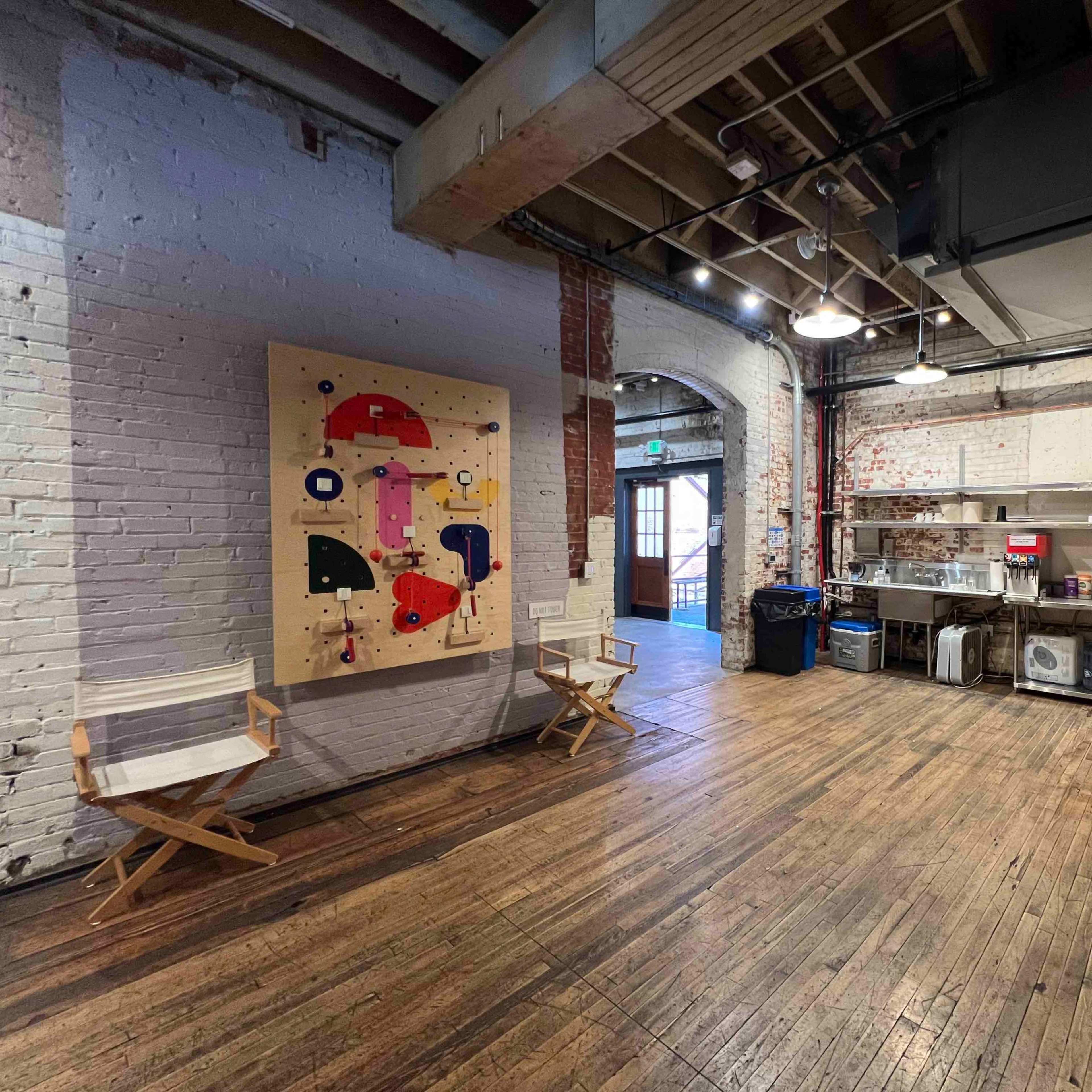 A spacious room with wooden floors, featuring a colorful abstract painting on the wall and a kitchen area with appliances in the background.