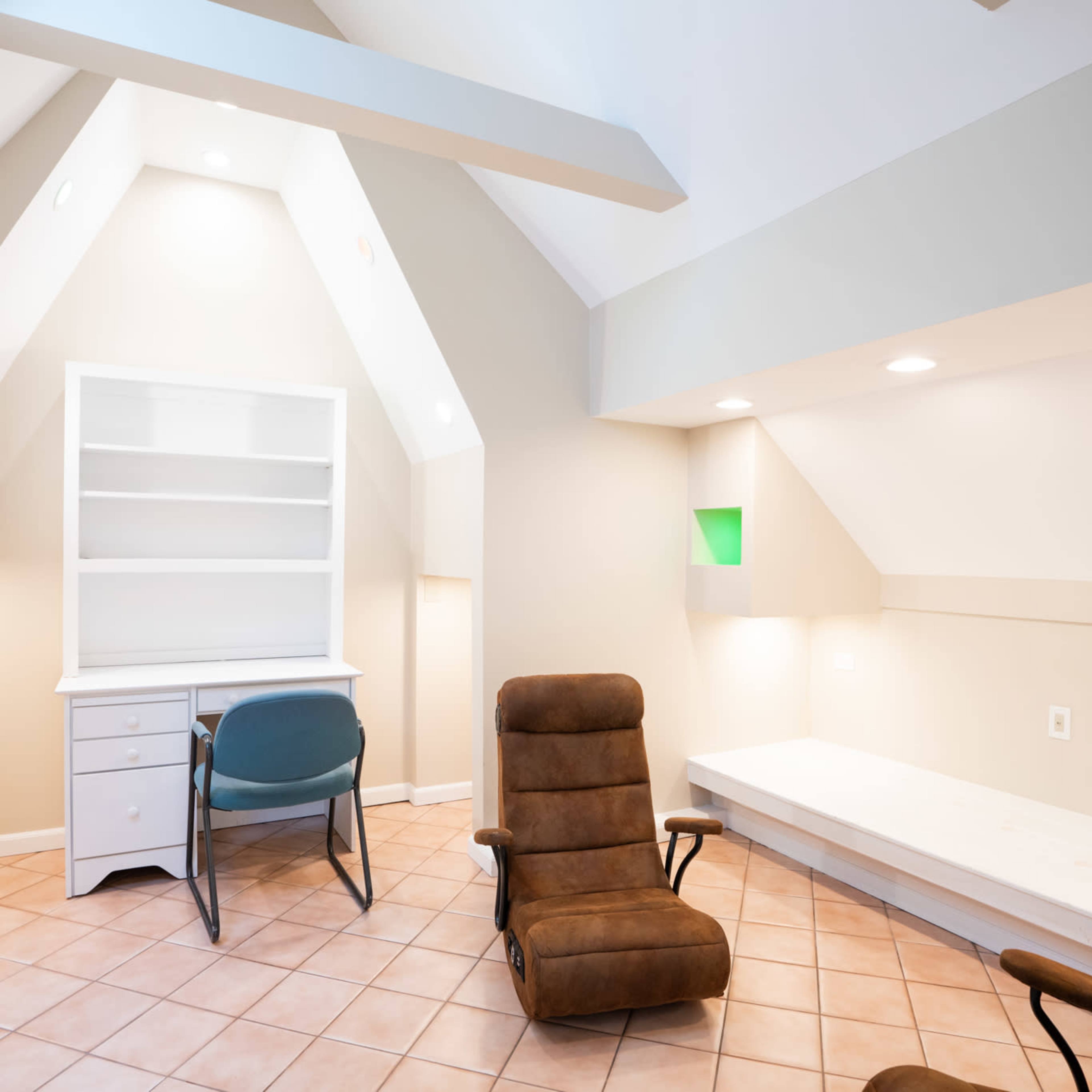 A small room with a sloped ceiling, featuring a desk, a chair, two reclining chairs, and a built-in bench along the wall.
