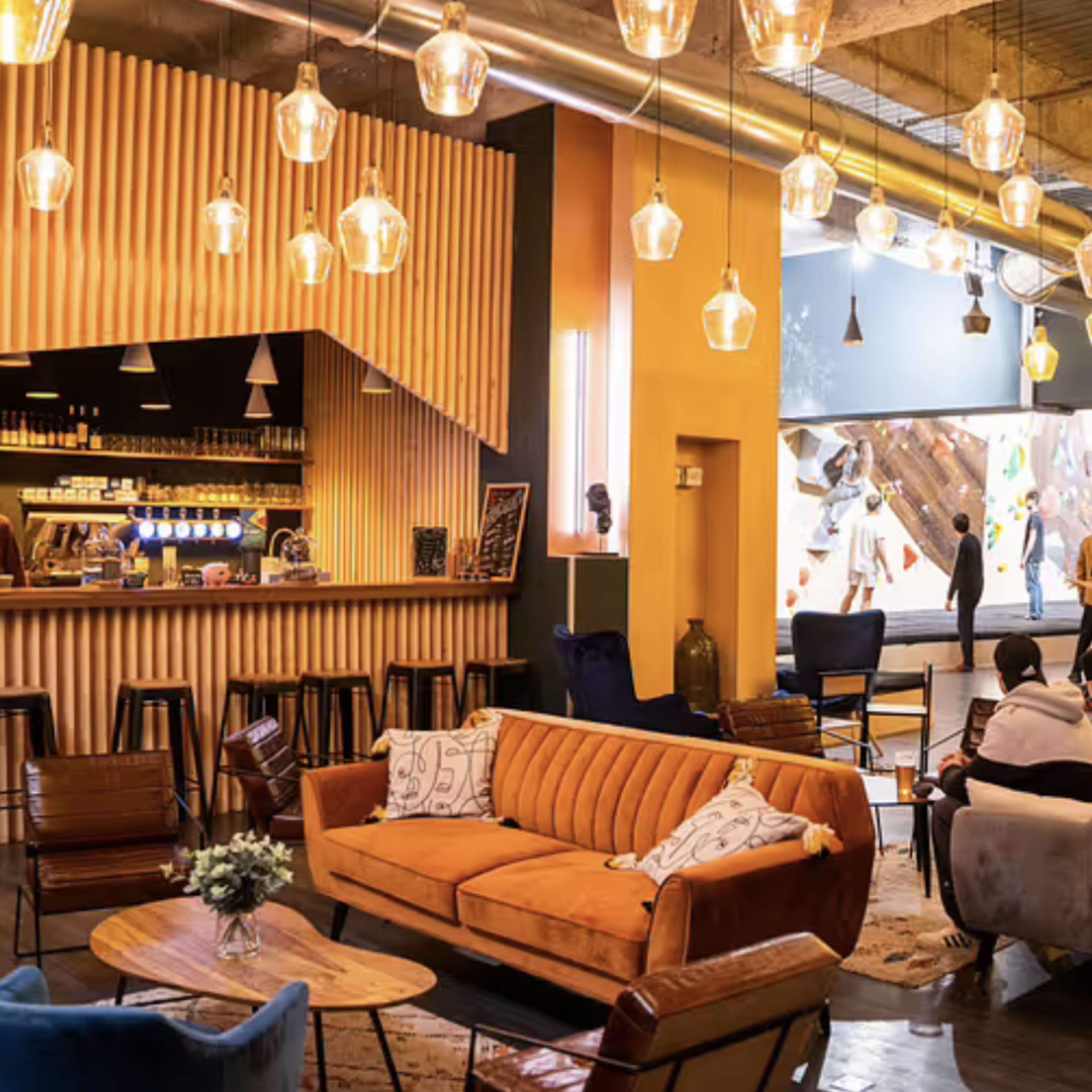 A vibrant indoor climbing gym with a café area featuring a wooden bar, a seating area with sofas, and climbing walls visible through large windows.