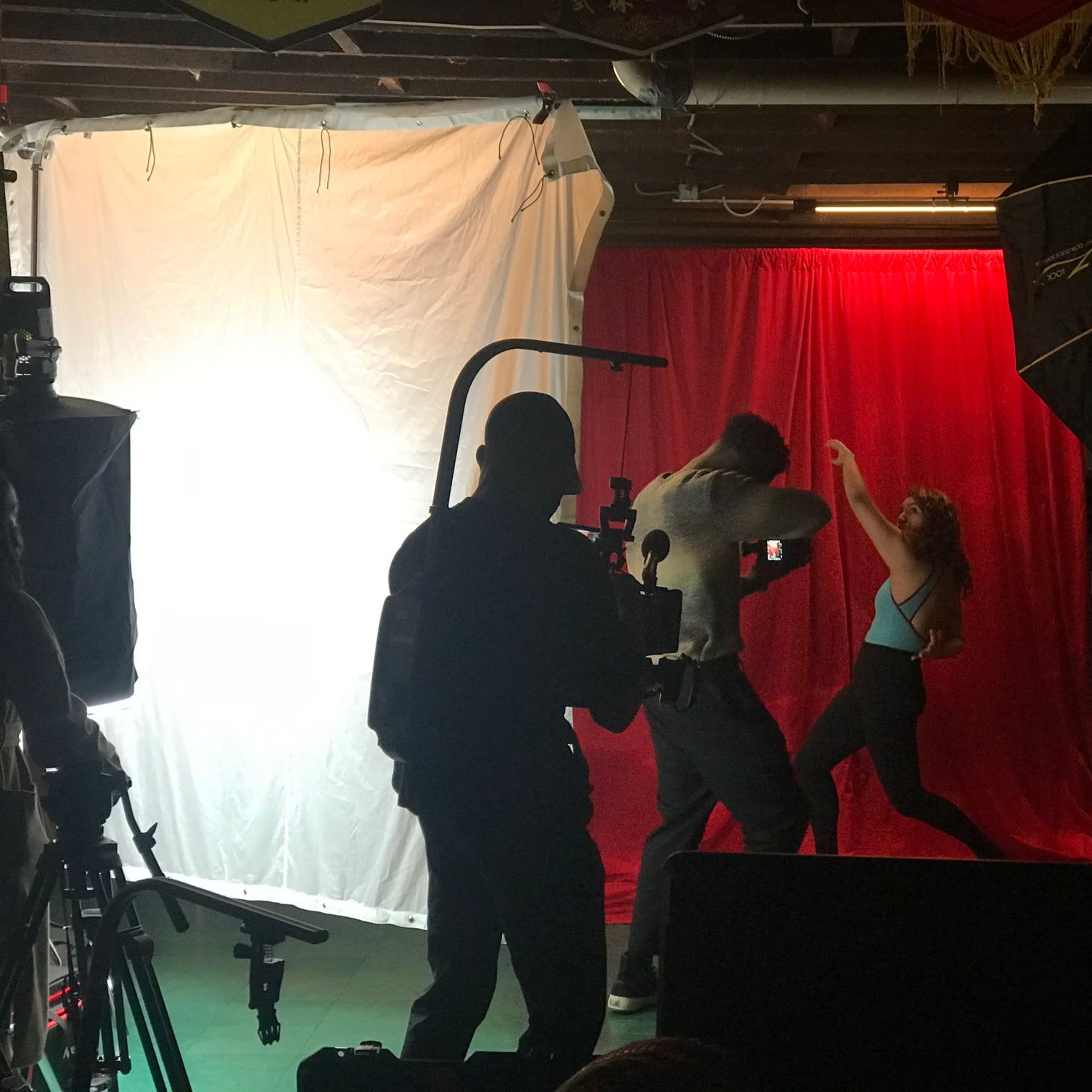 A photography shoot is in progress in a basement with a red backdrop, featuring a male model posing and a female model dancing, while a photographer captures the scene.