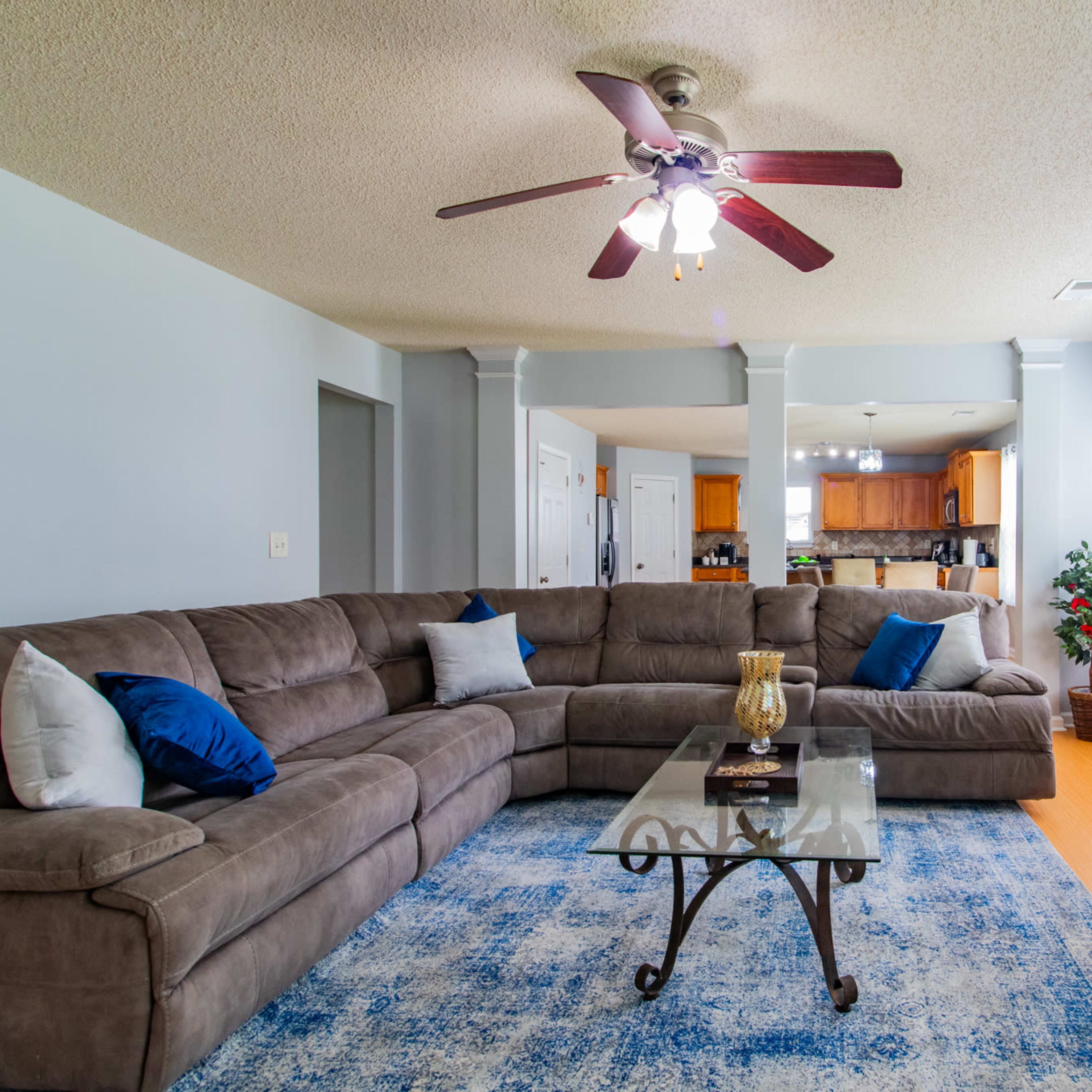 A spacious living room features a large sectional sofa, a glass coffee table, and a fireplace, with an open view into the kitchen area.