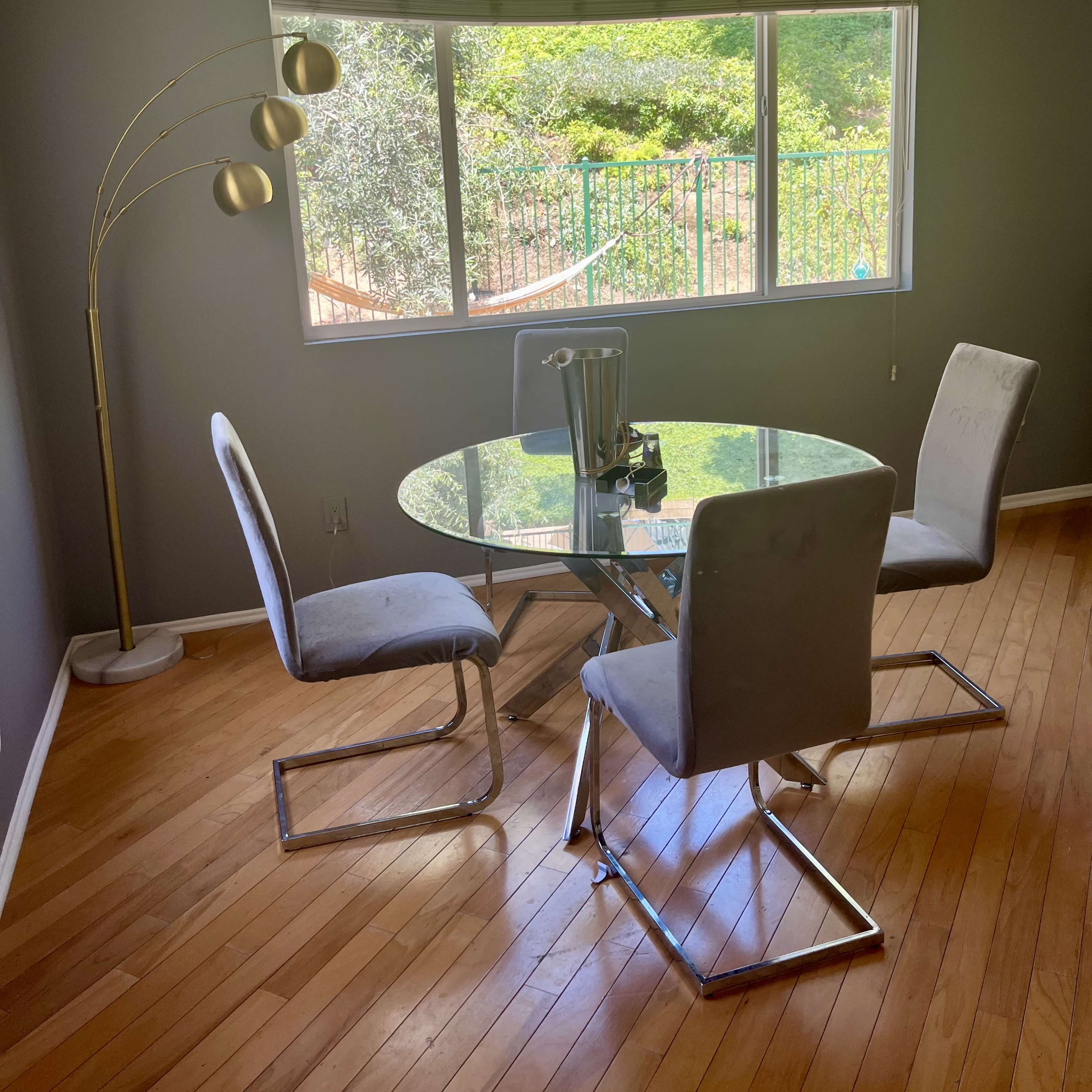A circular glass dining table is set with three chairs in a well-lit room, featuring a large window that overlooks a garden.