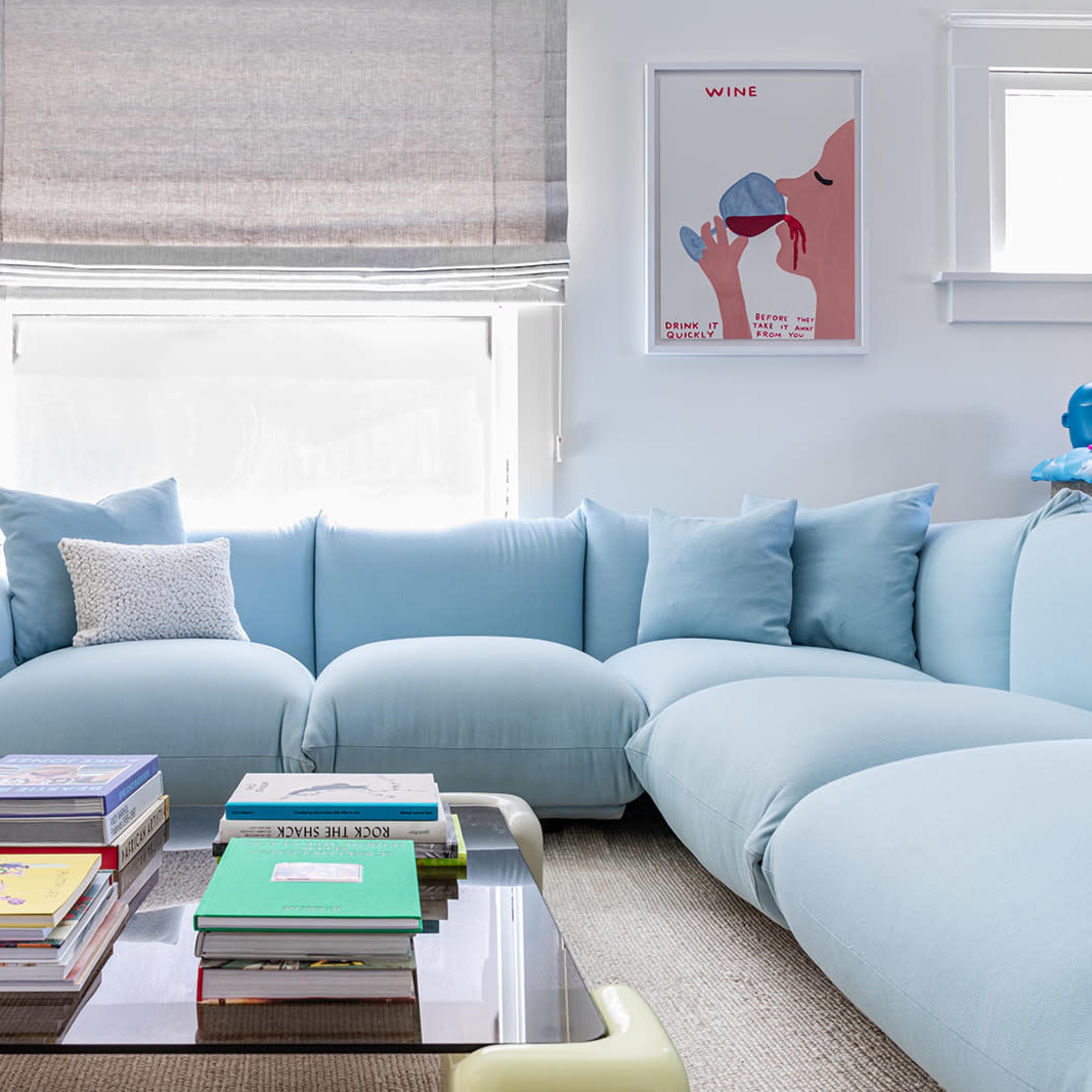 A light blue sectional sofa with a coffee table stacked with books, set against a backdrop of a window with shades and a framed poster on the wall.