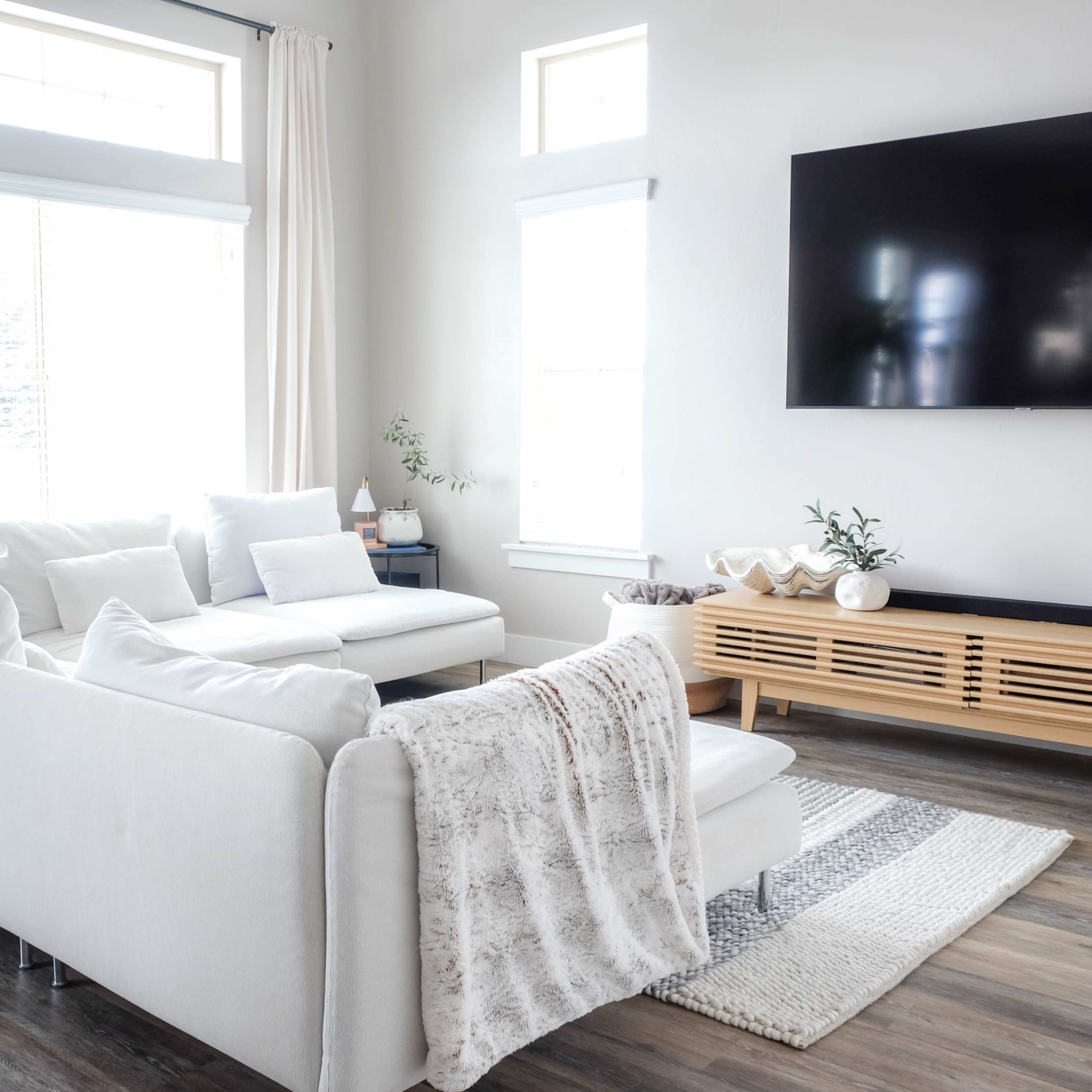 A bright living room features a white sectional sofa, a wooden media console, and large windows letting in natural light.