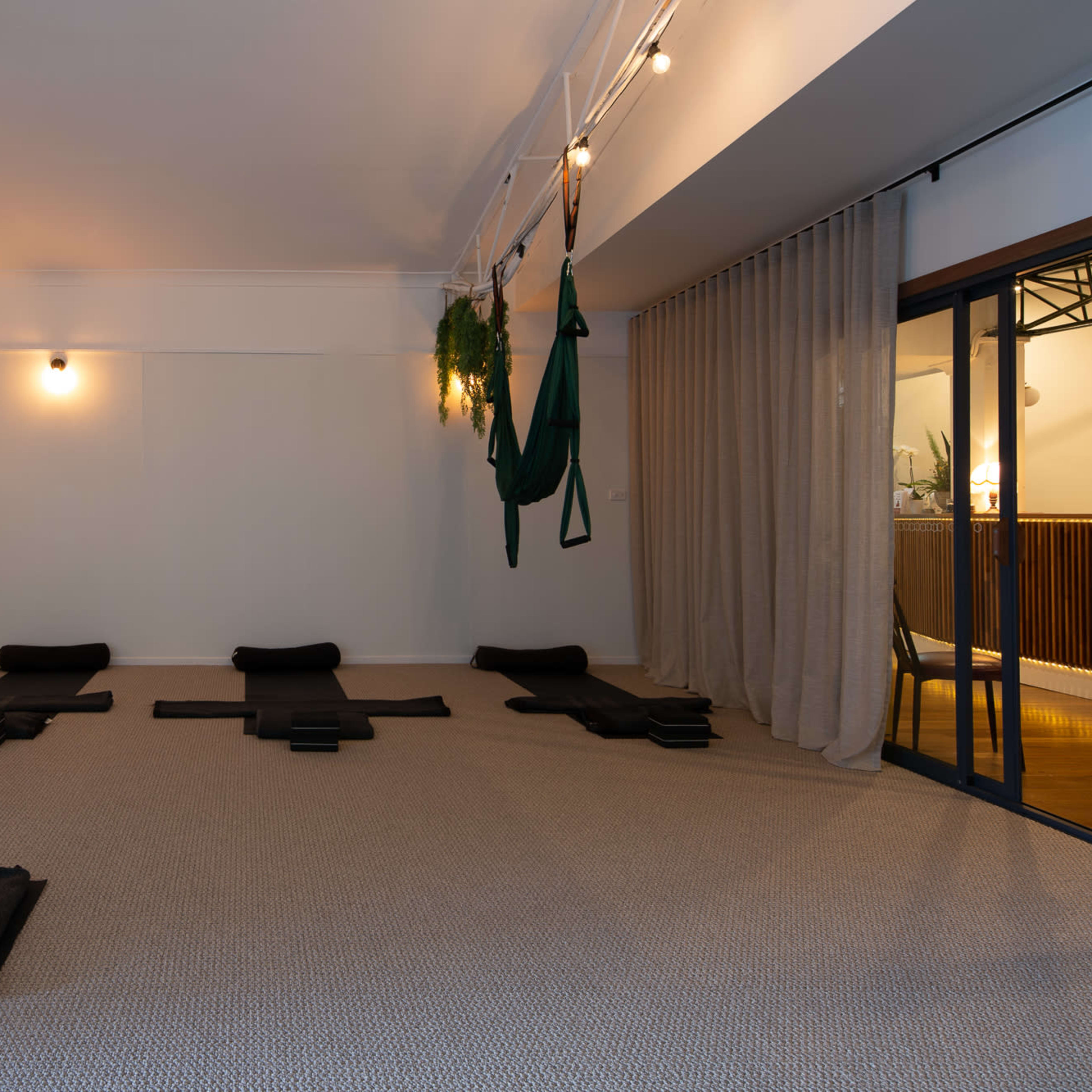 A spacious yoga studio with several black mats arranged on a carpeted floor, a large mirror on one wall, and curtains partially separating the space.