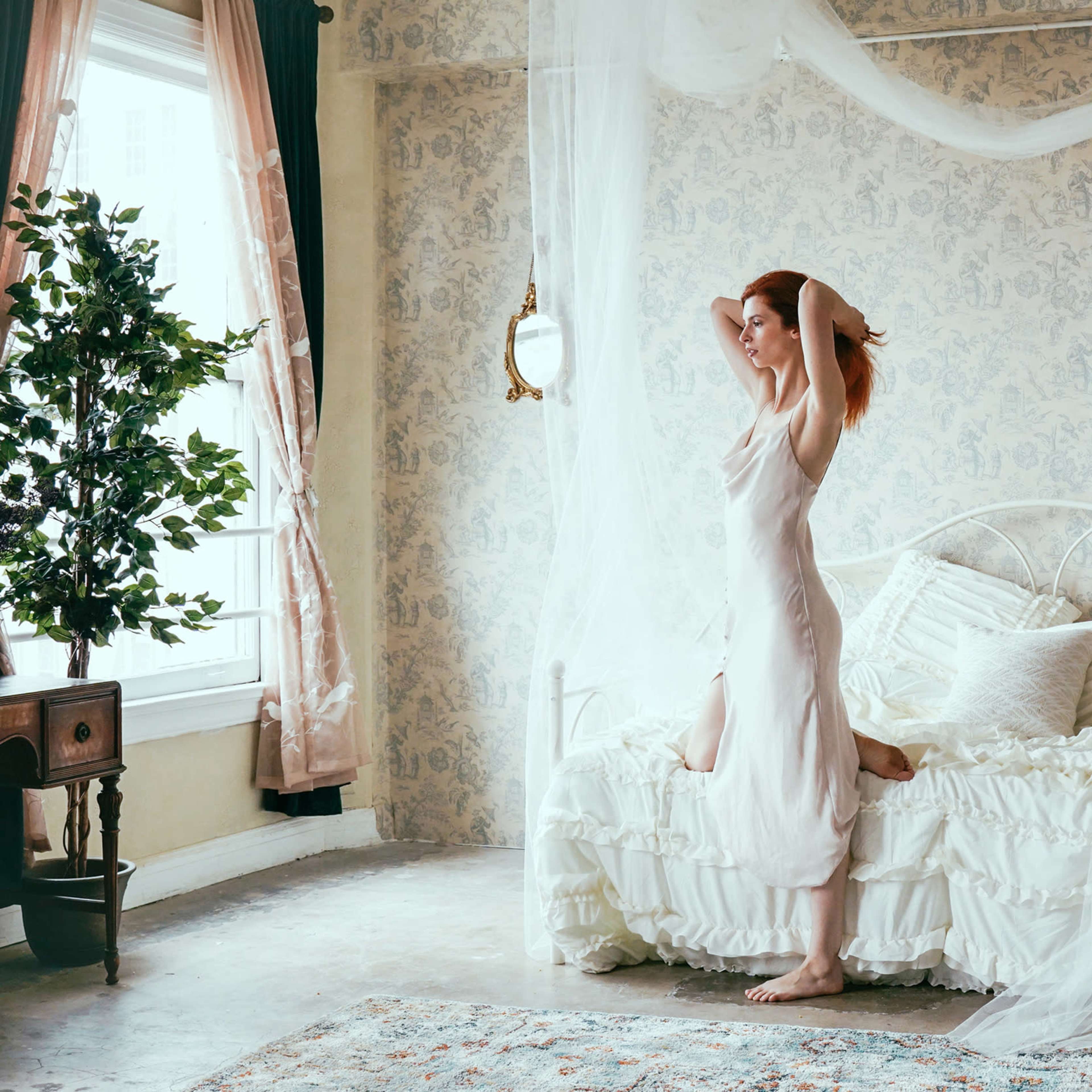 A woman stands on a bed in a softly lit room, surrounded by sheer curtains and vintage decor.