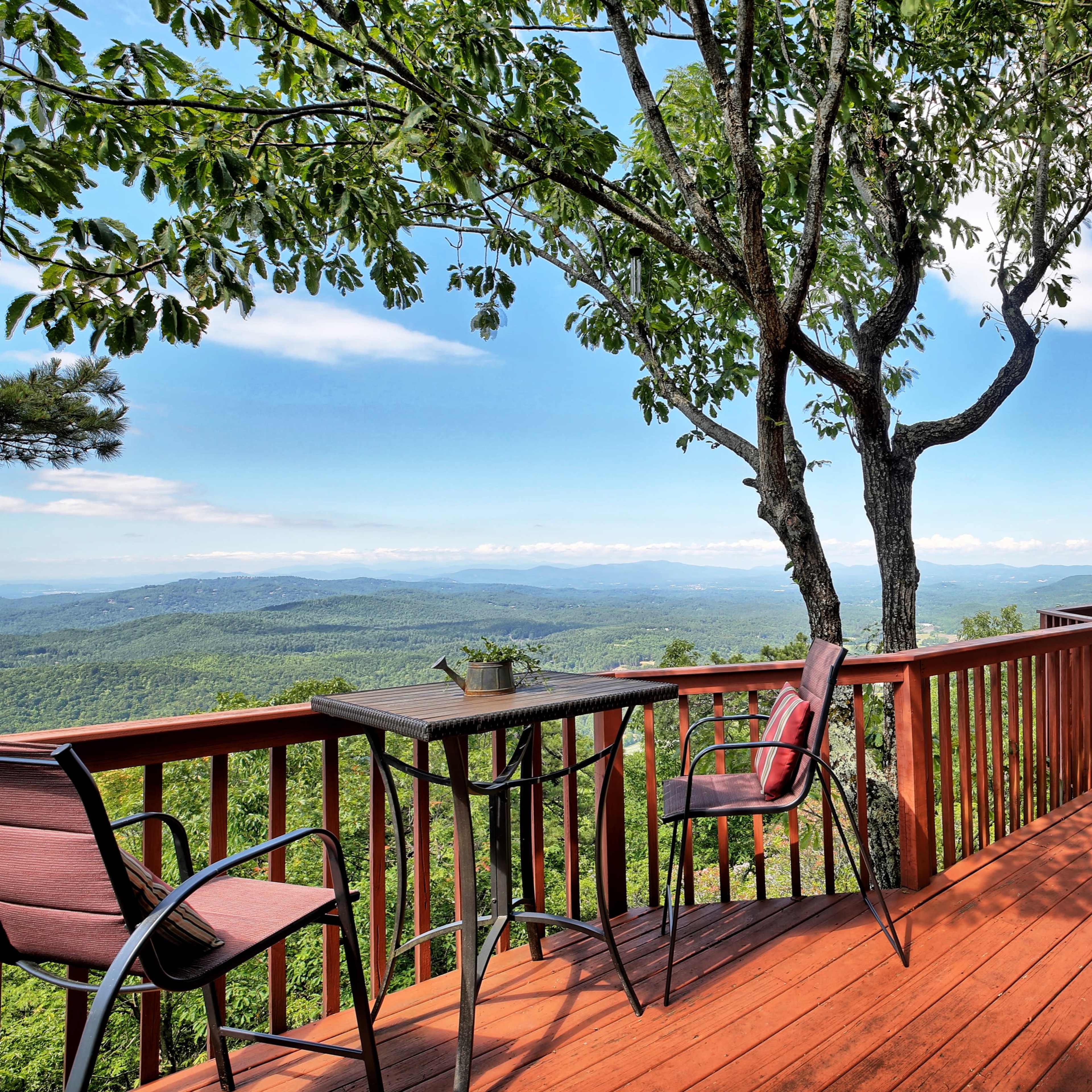 A wooden deck overlooking a mountainous landscape with a table and chairs near a tree.