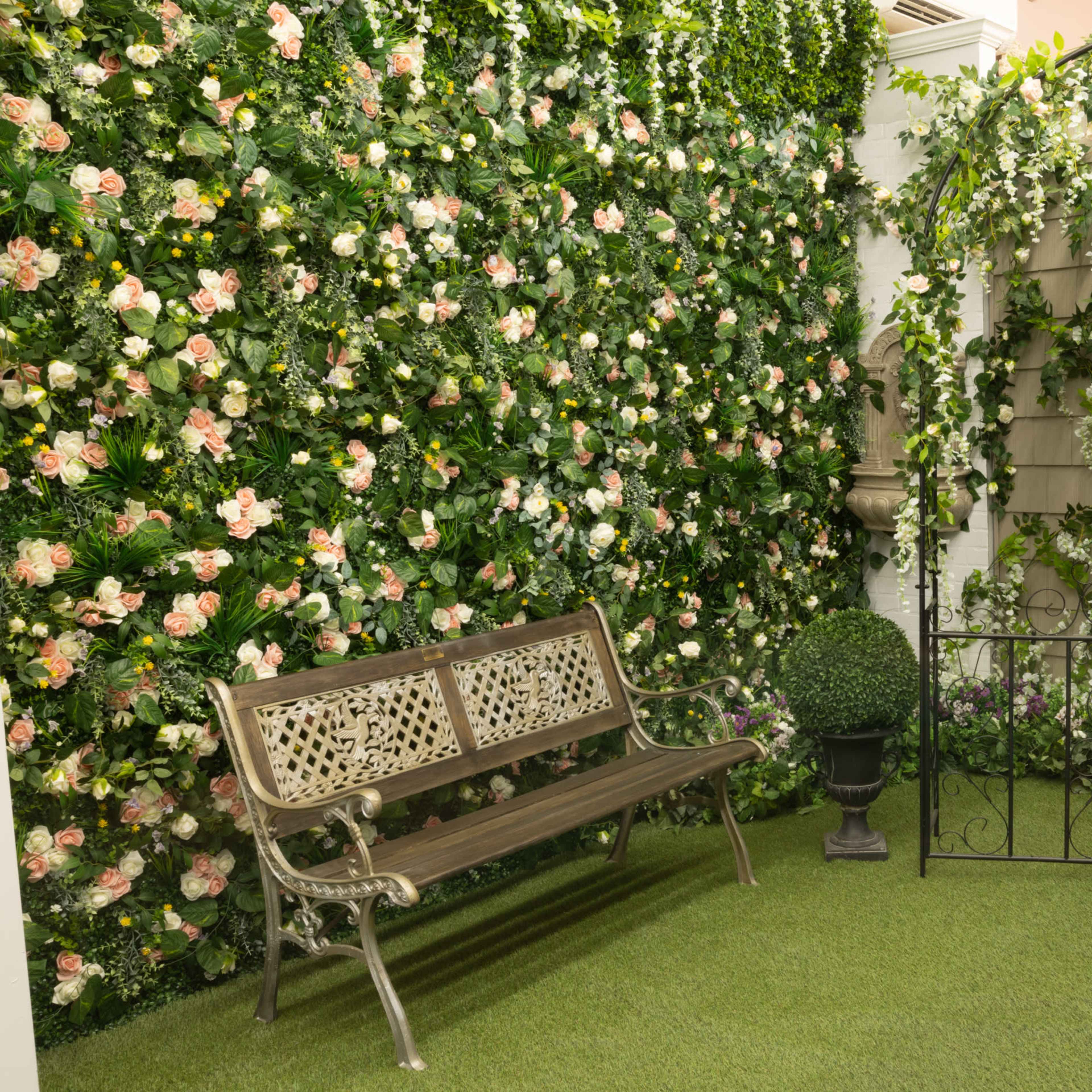 A decorative garden corner featuring a bench, a flower-covered wall, a window, and a small gate with potted plants.
