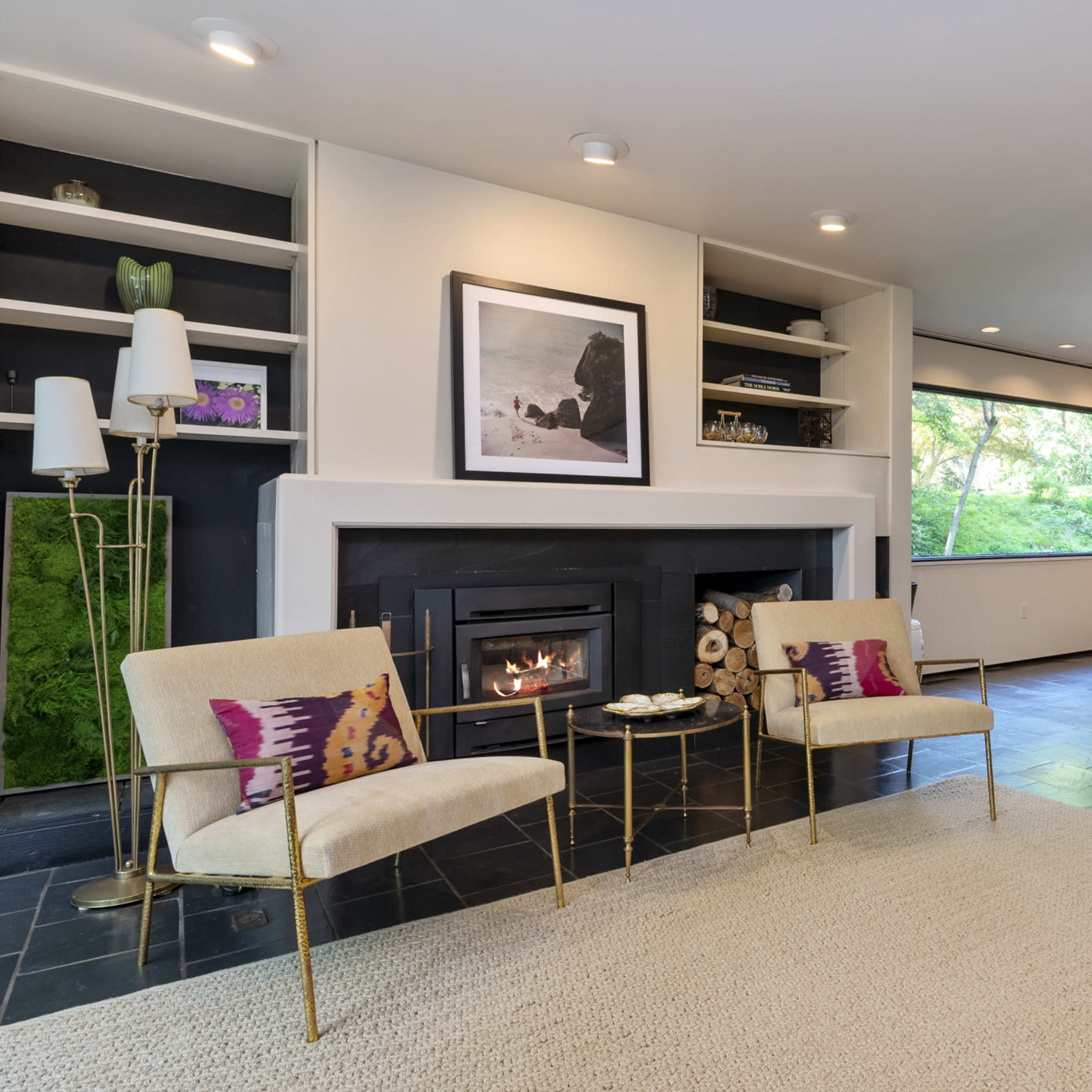 A modern living room with a fireplace, two chairs, a rug, and a view of greenery through a large window.