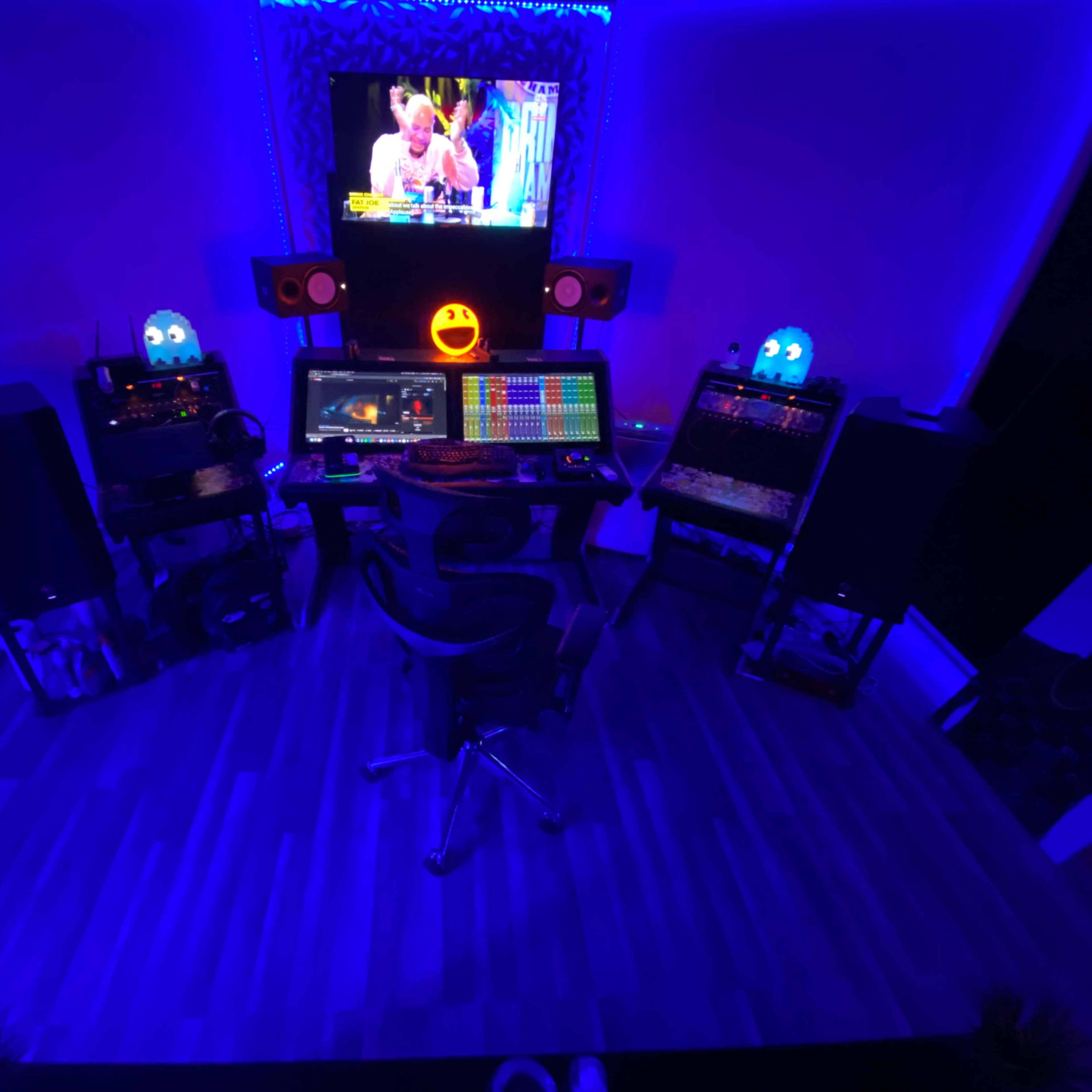 A modern music production studio illuminated with blue lights, featuring a central workstation with multiple screens, audio equipment, and speakers.