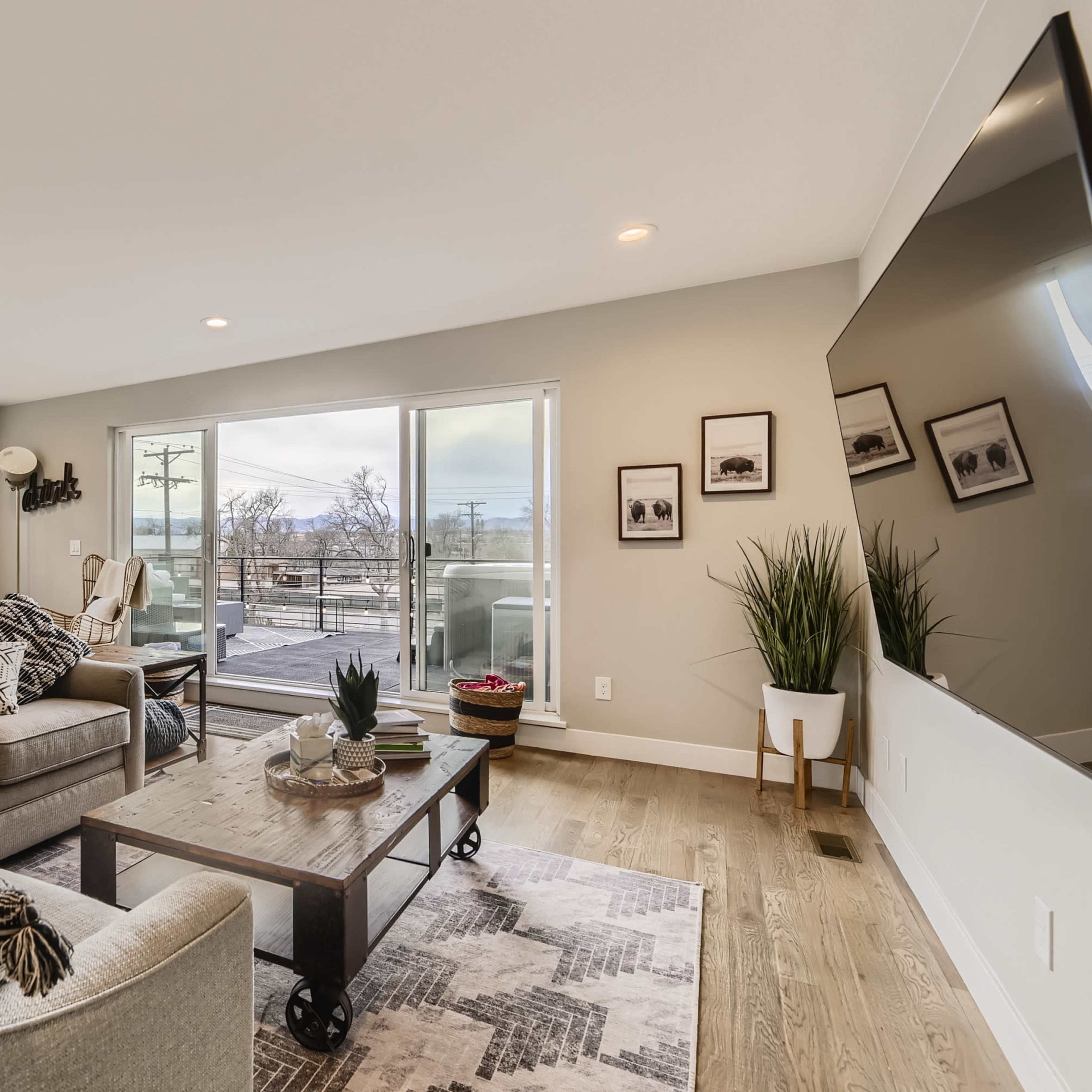 The living room features a gray sectional sofa, a wooden coffee table, and large sliding glass doors that open to a balcony with outdoor seating.