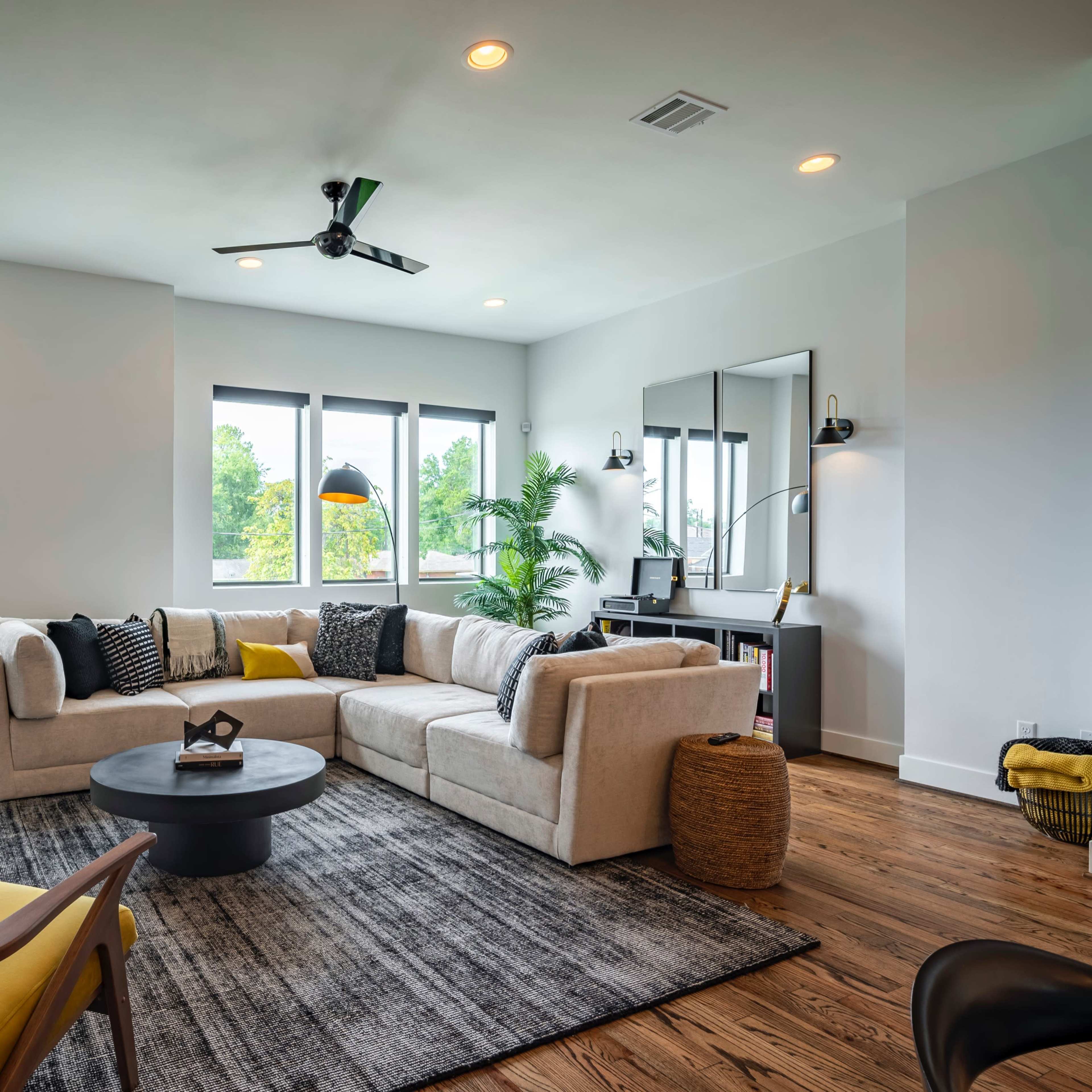 A modern living room features a sectional sofa, a round coffee table, and large windows that provide natural light.