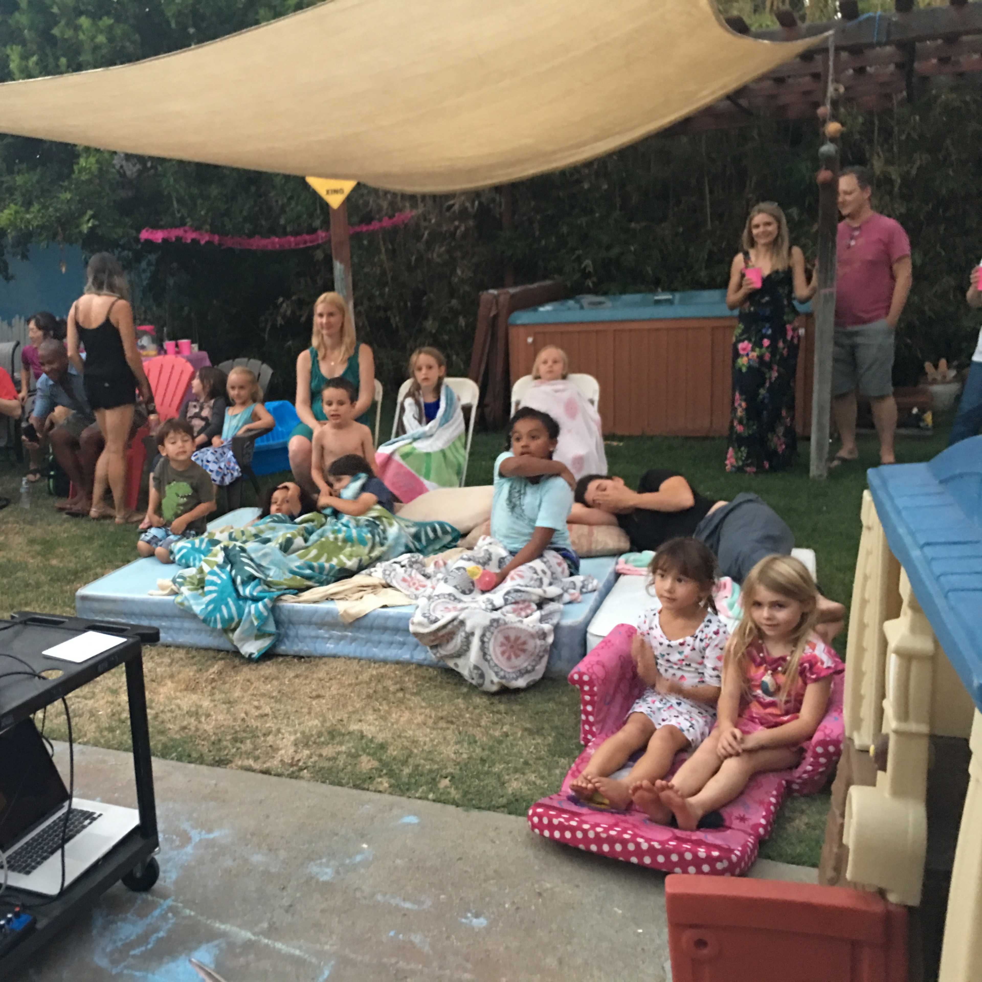 A group of children and adults gather in a backyard setting, seated on blankets and in chairs, while a projector displays an image onto a screen.