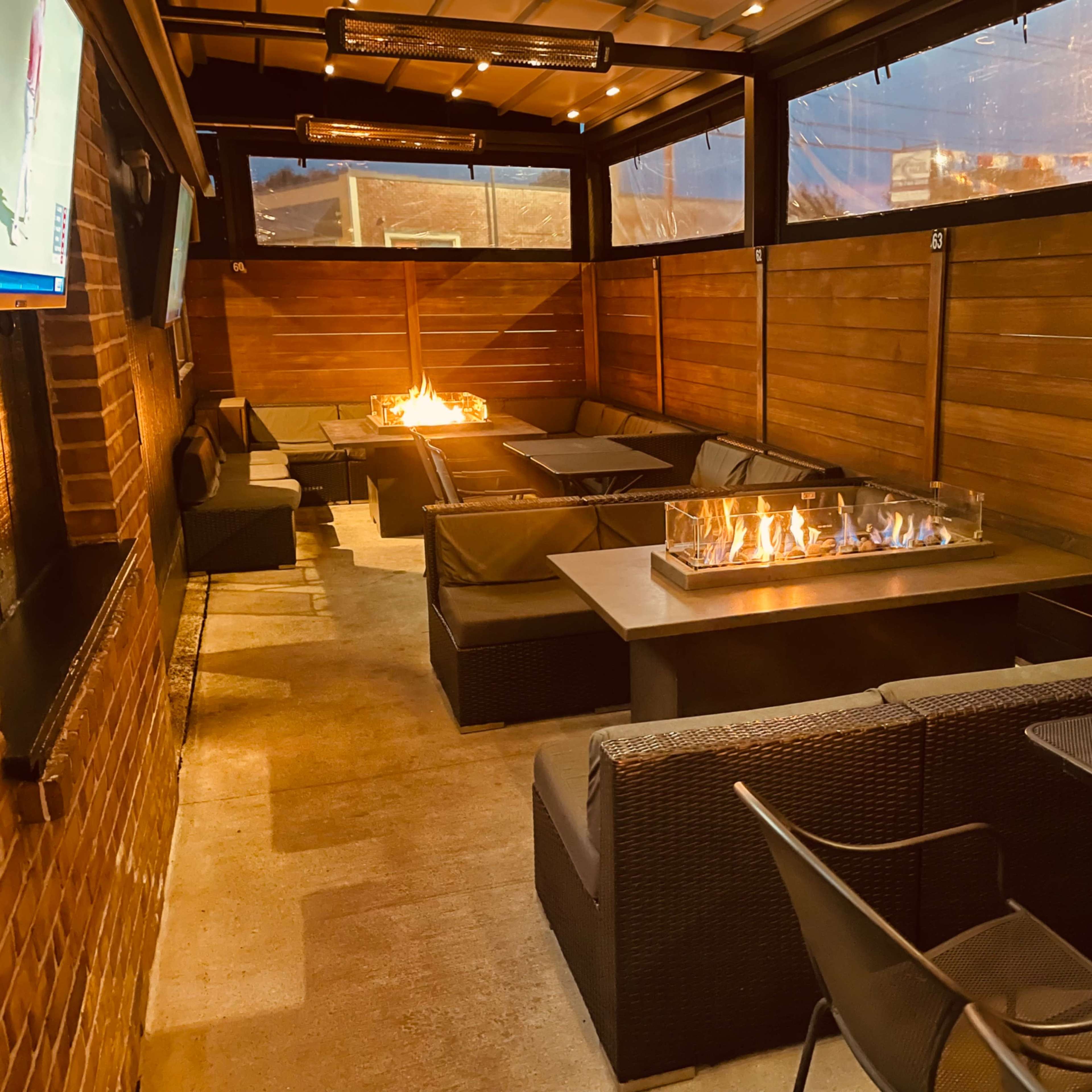 A cozy outdoor seating area with dark wicker furniture and a gas fire pit in the center, enclosed by wooden walls and a roof.