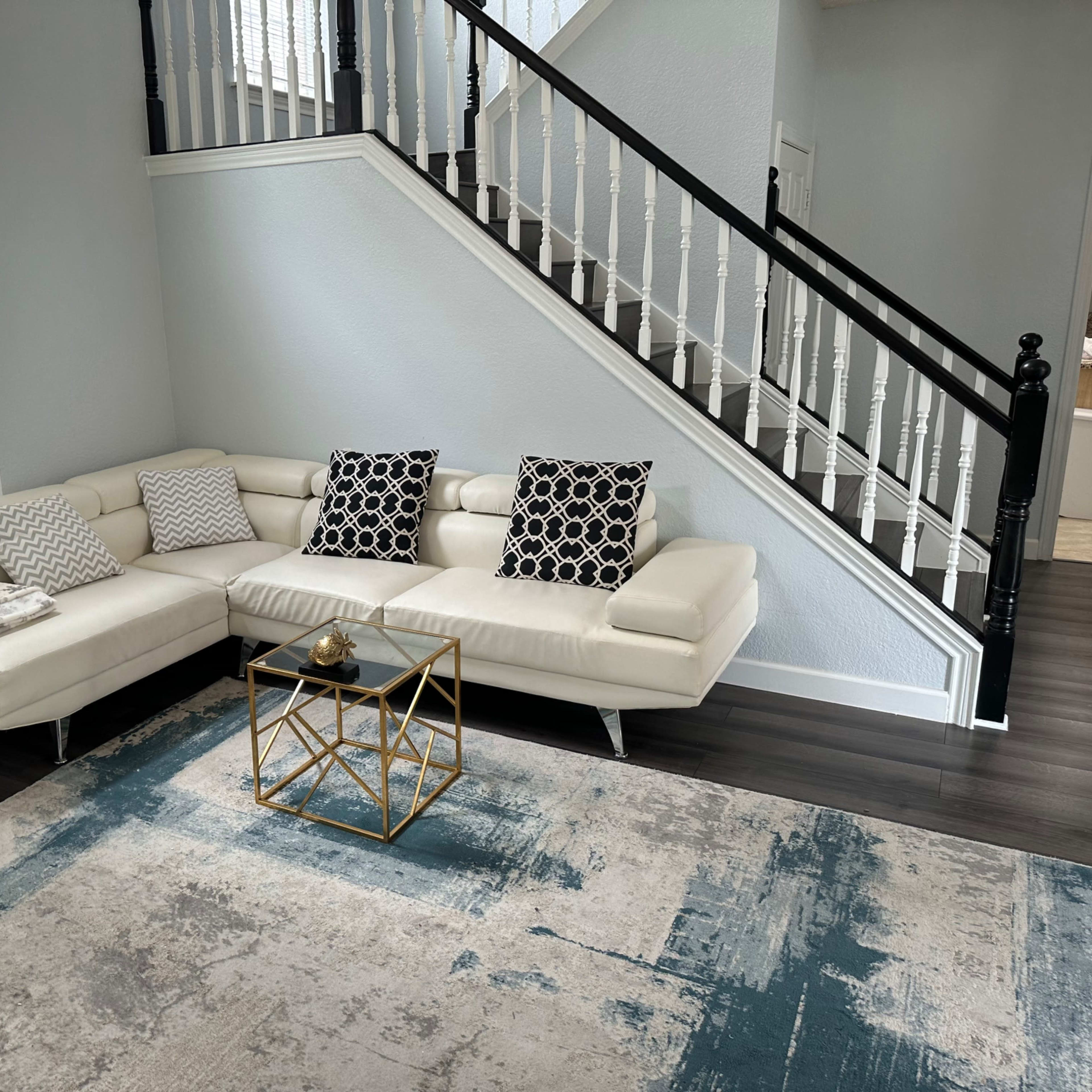 A modern living room featuring a white sectional sofa with patterned pillows, a gold geometric coffee table, and a textured area rug, set against a backdrop of a staircase with black railing.