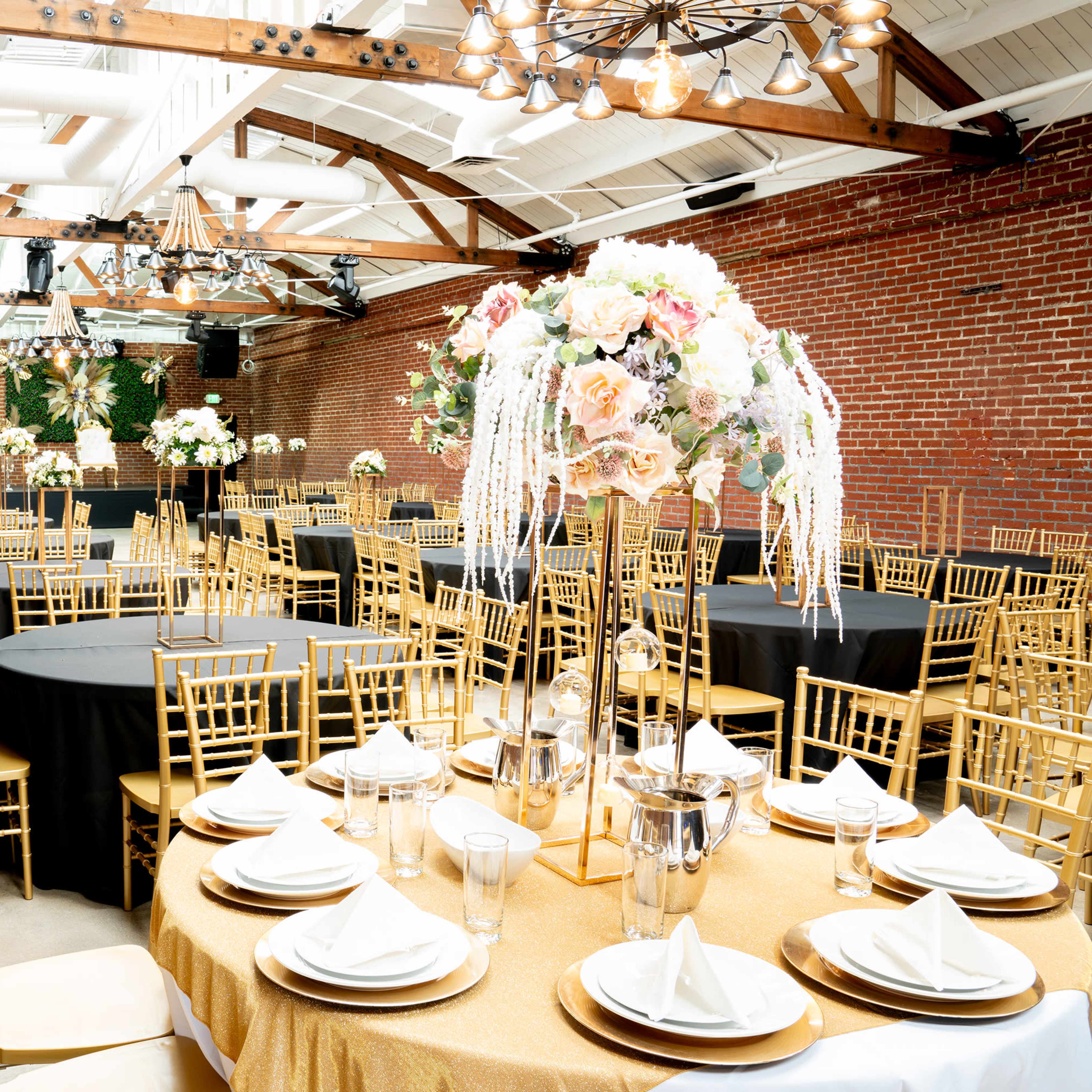A spacious event venue features round tables set with elegant tableware and decorative floral centerpieces, surrounded by gold chiavari chairs.