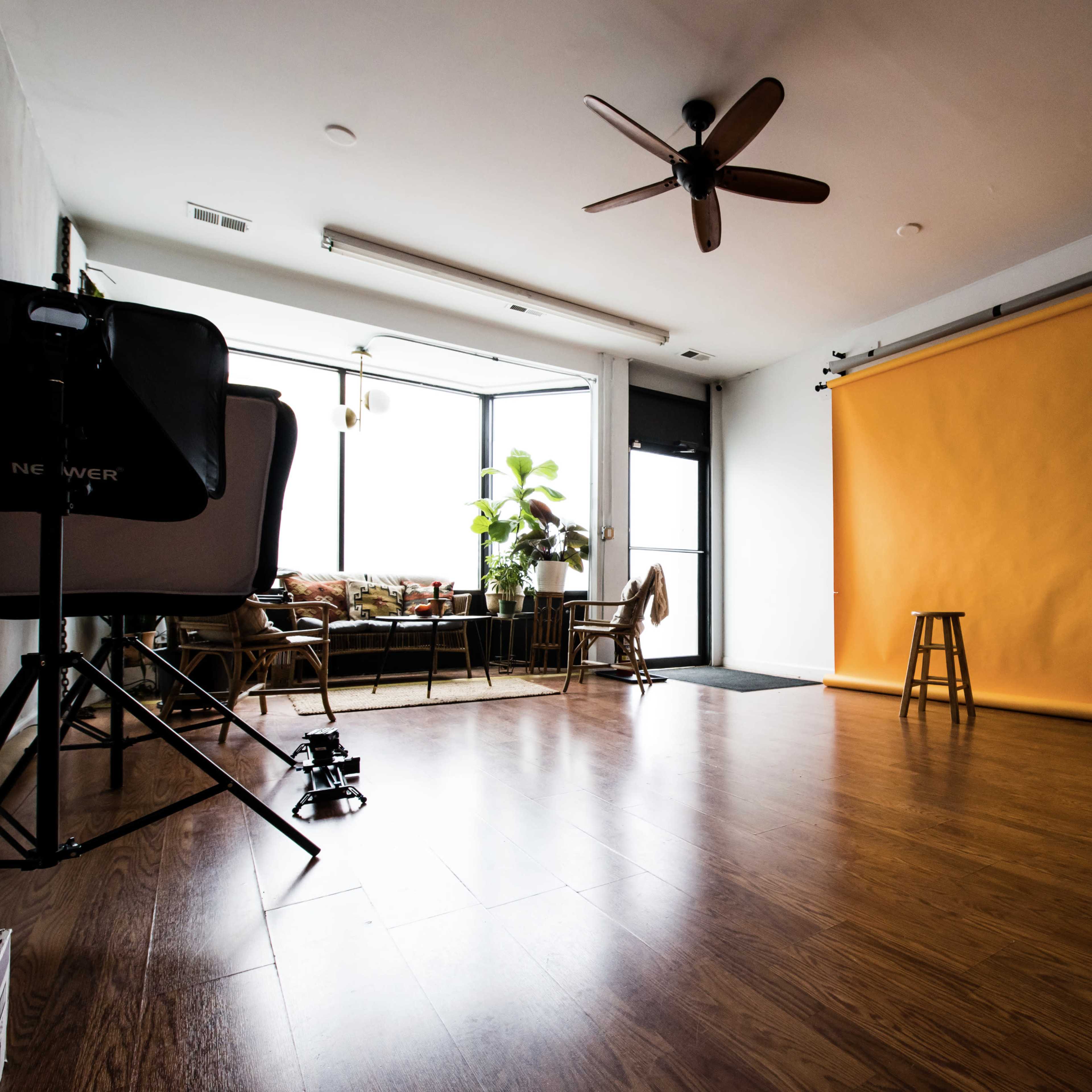 A photography studio features a yellow backdrop, wooden flooring, and a seating area with plants in the background.