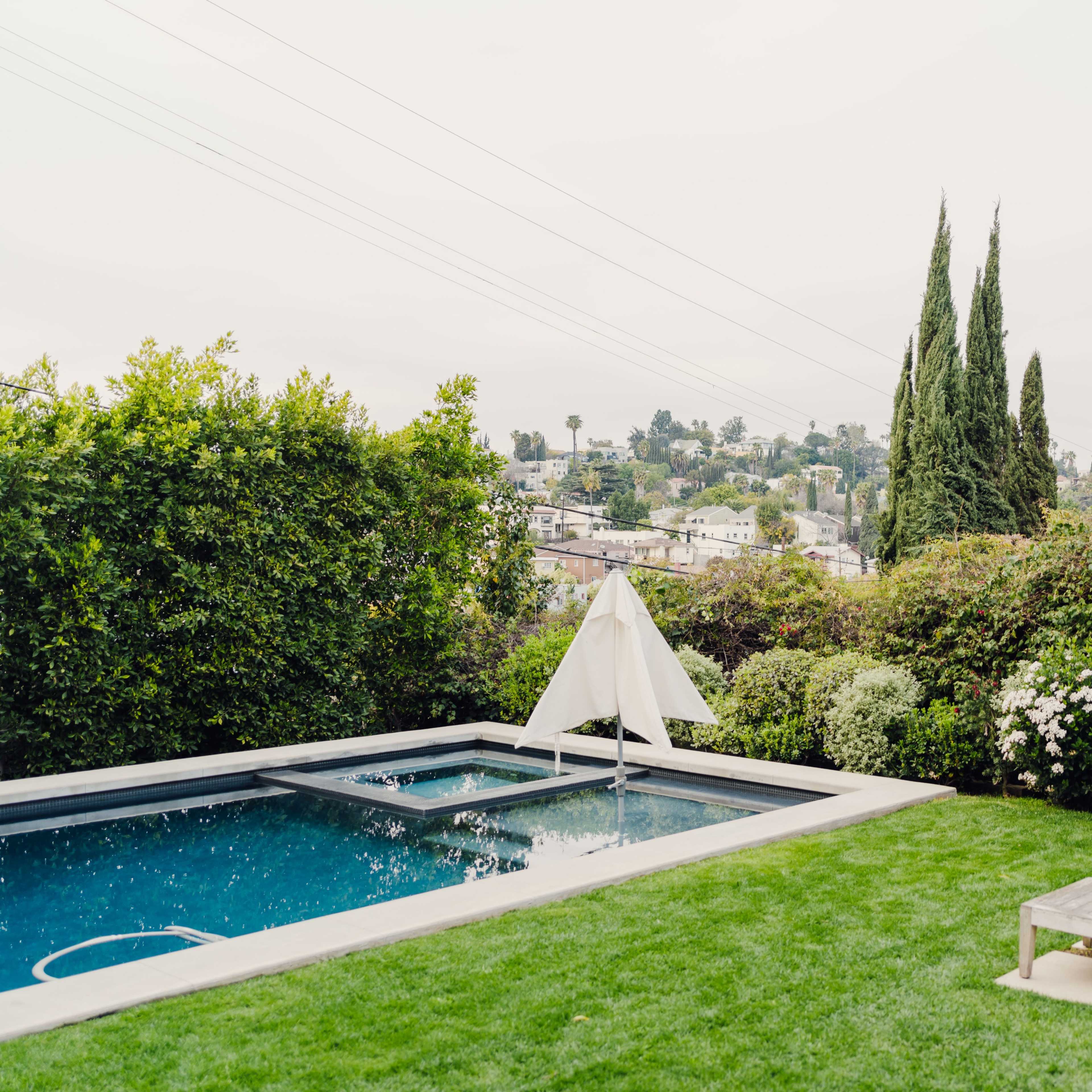 A rectangular swimming pool is located in a landscaped backyard with greenery, a small wooden table and chairs, and a view of houses and trees in the distance.