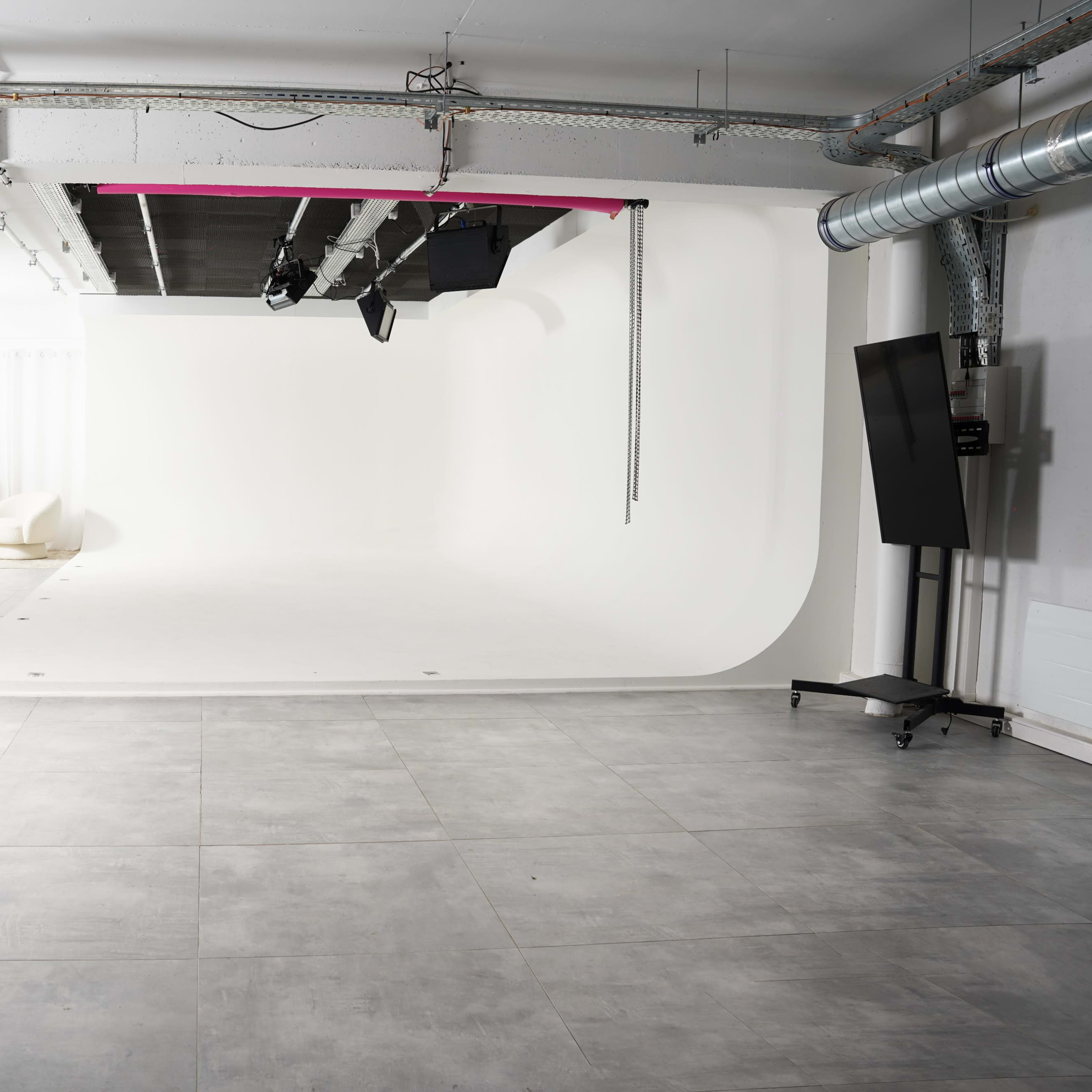 A spacious photography studio with a seamless white backdrop and a light setup, featuring grey tiled flooring and modern furniture in the corner.