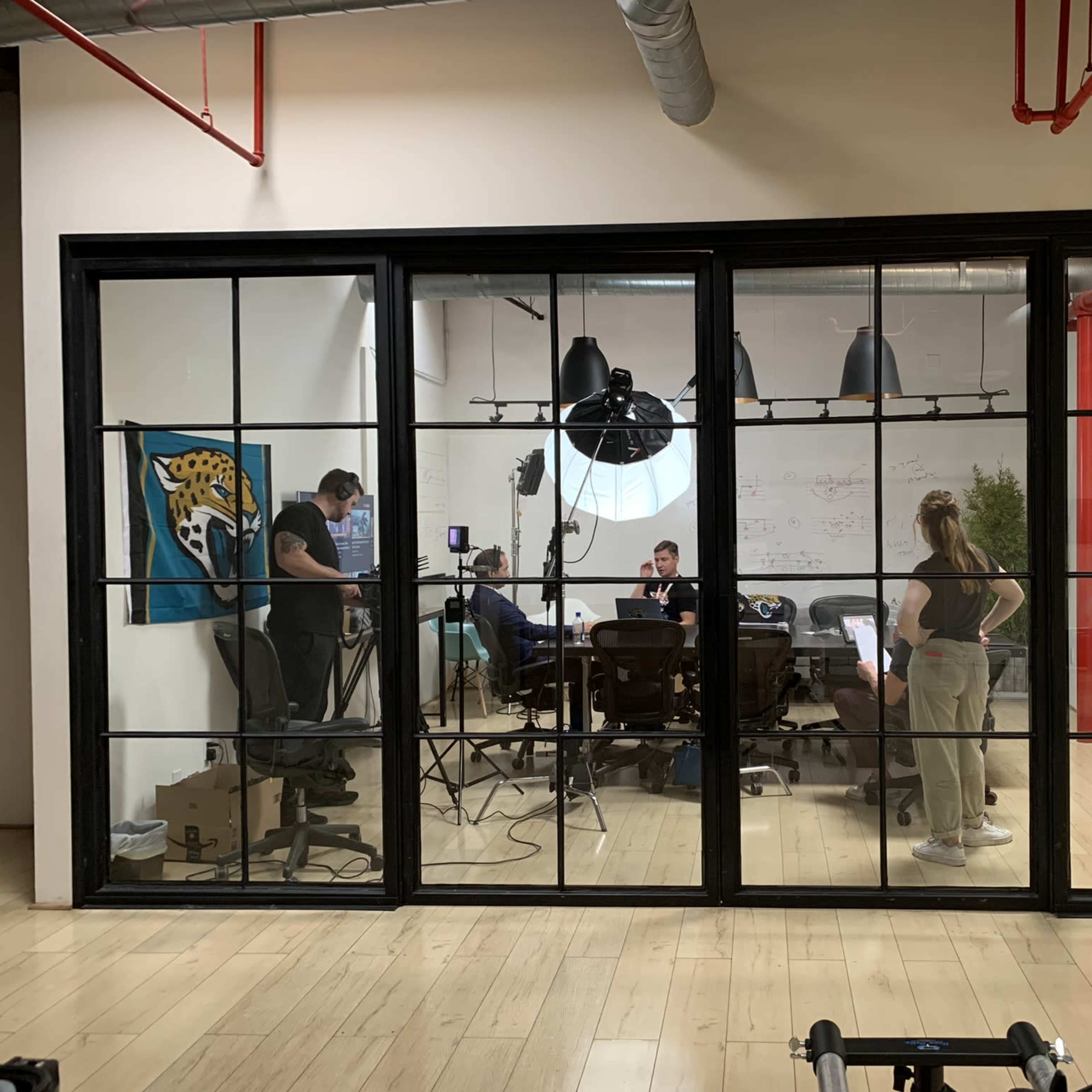 A group of people is working in a modern conference room, visible through large windows, with studio lighting equipment set up for a shoot.