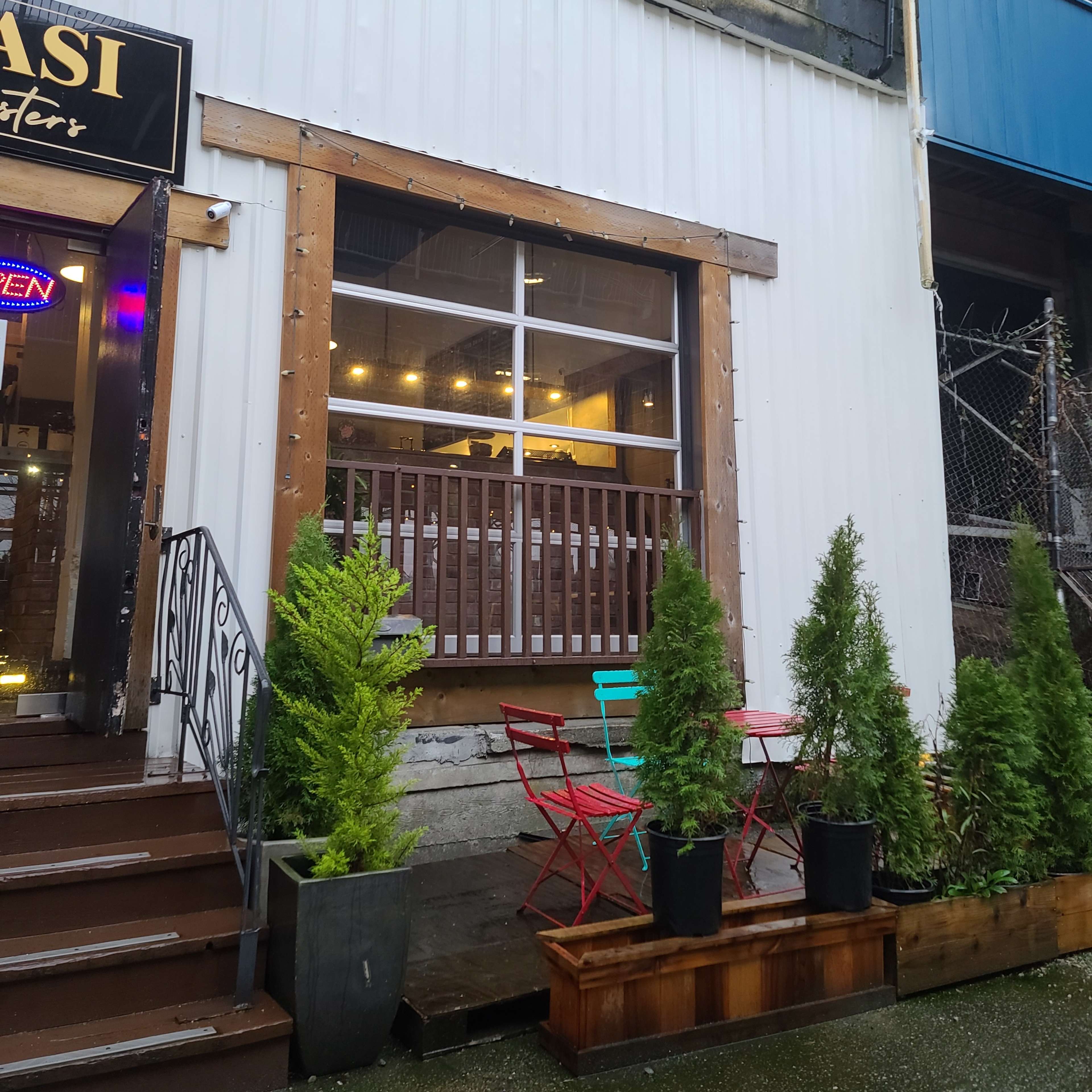 A café entrance with a wooden frame, flanked by two small evergreen shrubs, and featuring colorful outdoor seating with red and blue chairs.