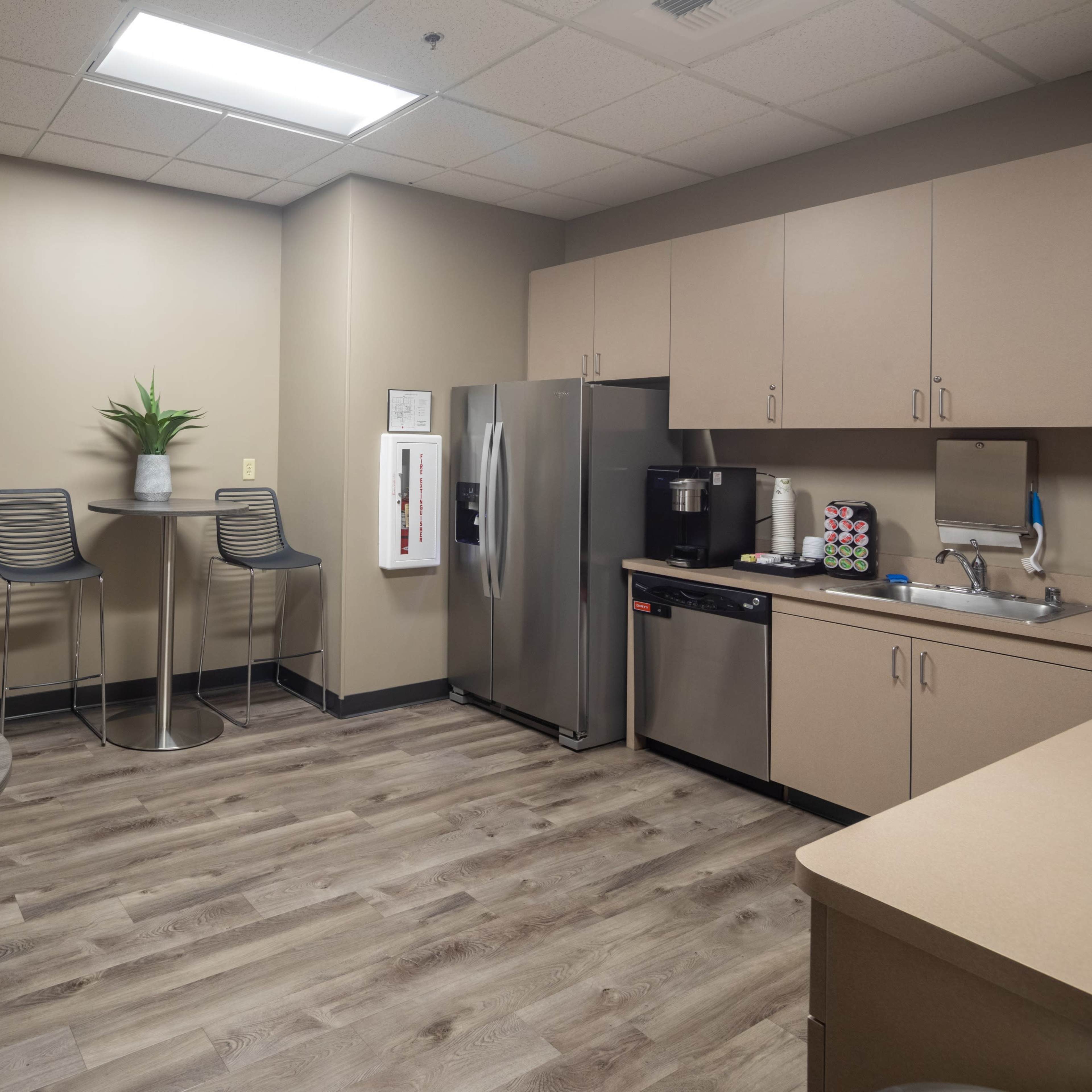 A modern kitchenette equipped with stainless steel appliances, a round table, two high chairs, and plants arranged on the countertop.