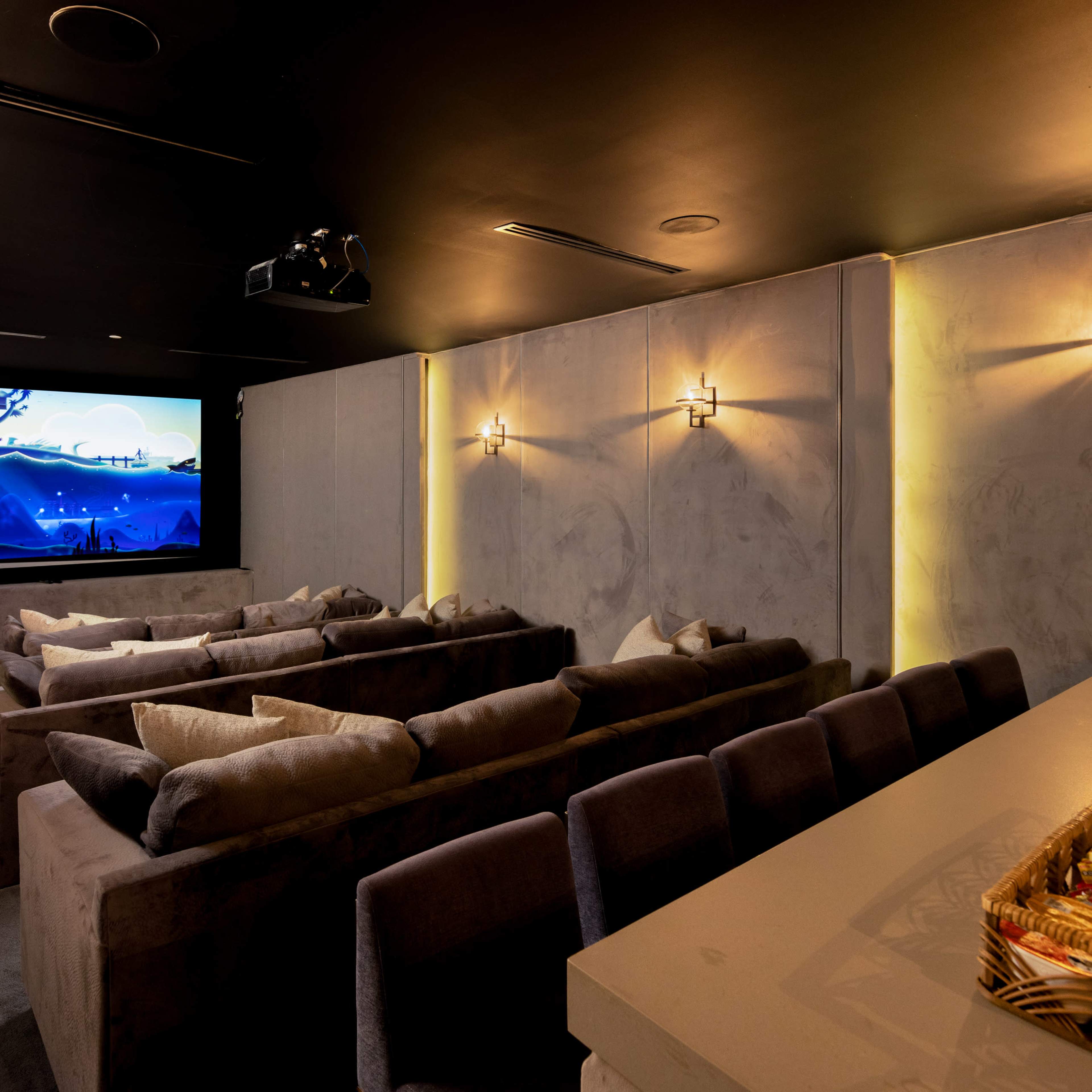 A home theater room with several rows of plush seating facing a large screen, illuminated by wall sconces and a snack table in the foreground.