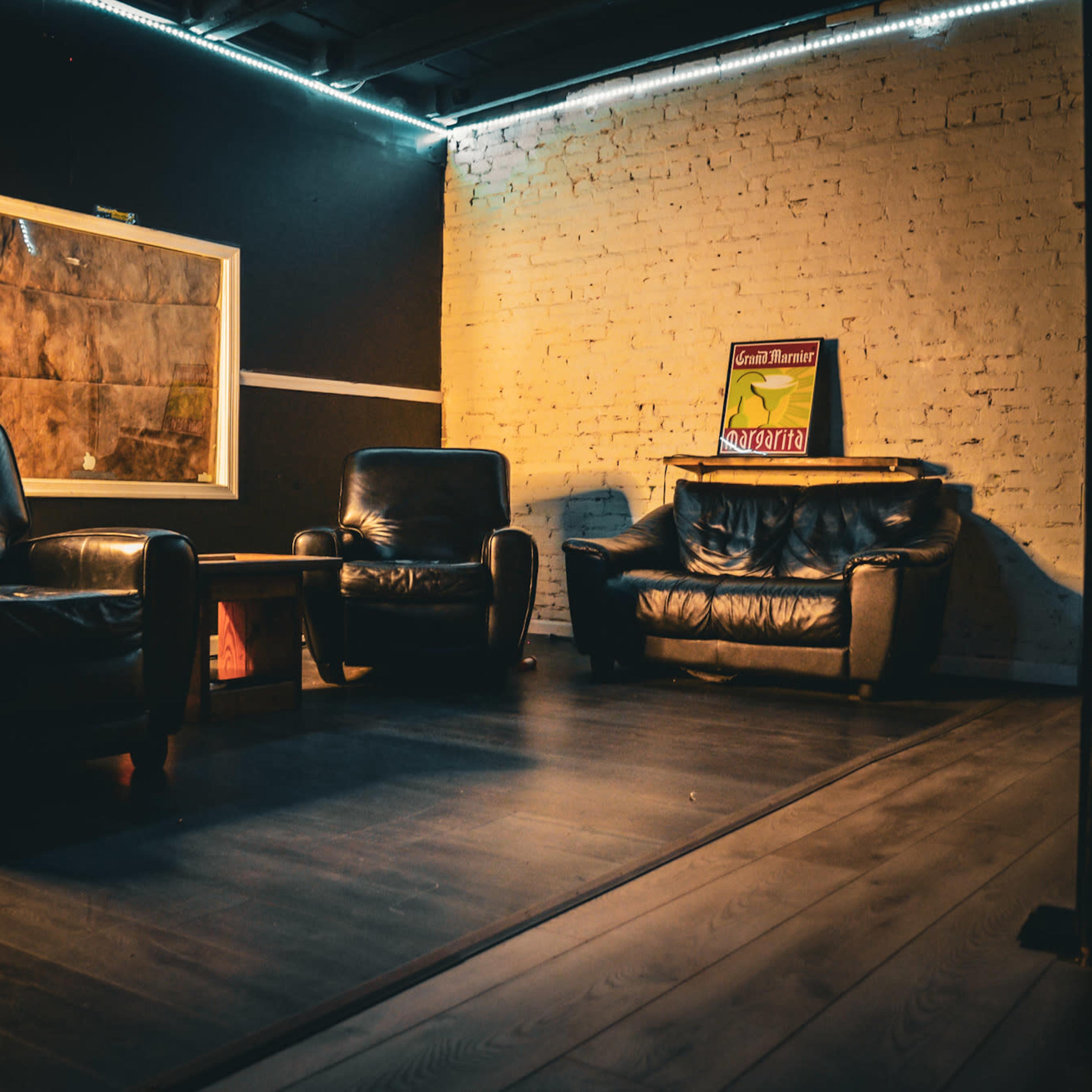 A dimly lit room features two black leather armchairs and a brown sofa against a white brick wall, with a framed map and a sign on display.