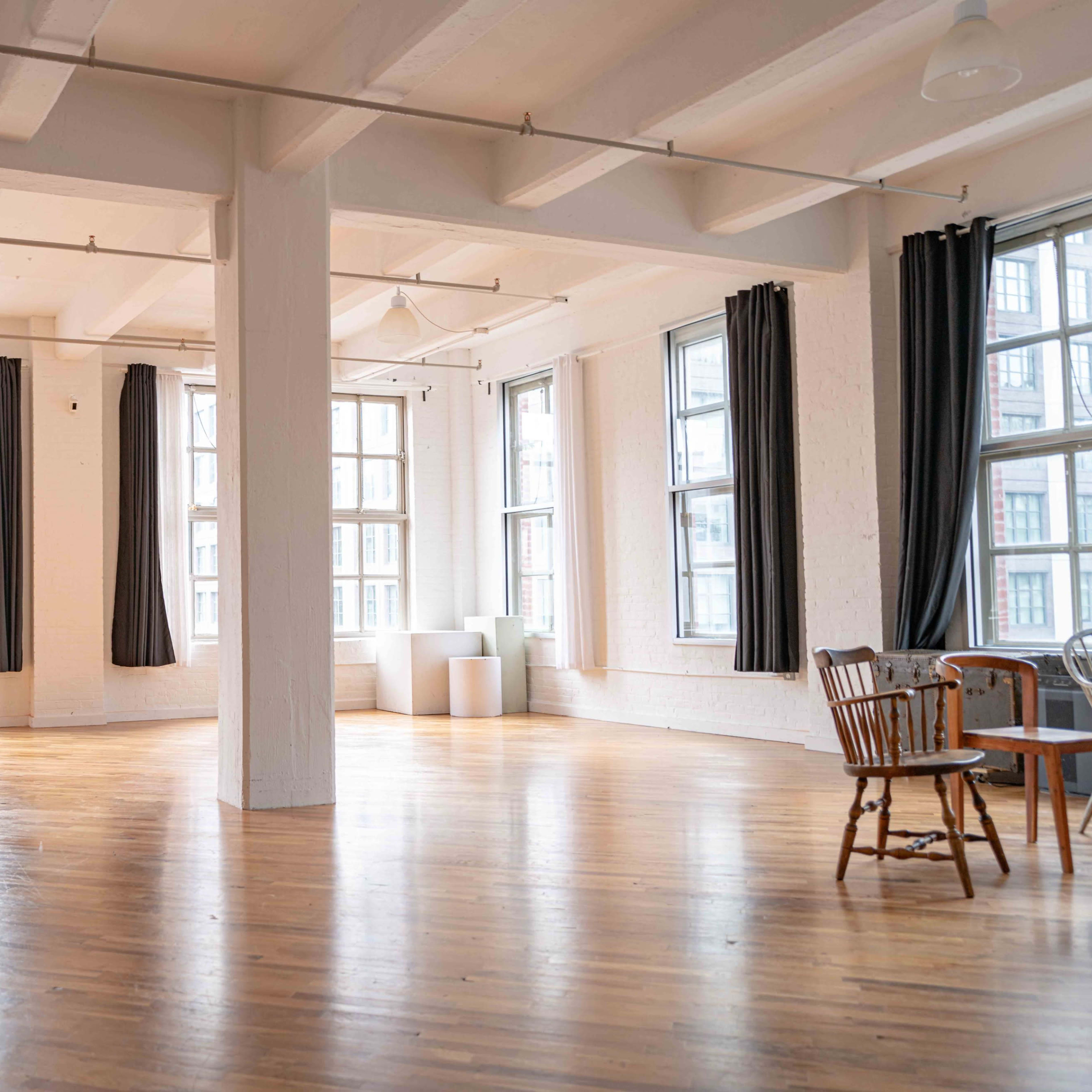 A spacious, well-lit room features large windows and wooden flooring, with a table and chairs near the corner.