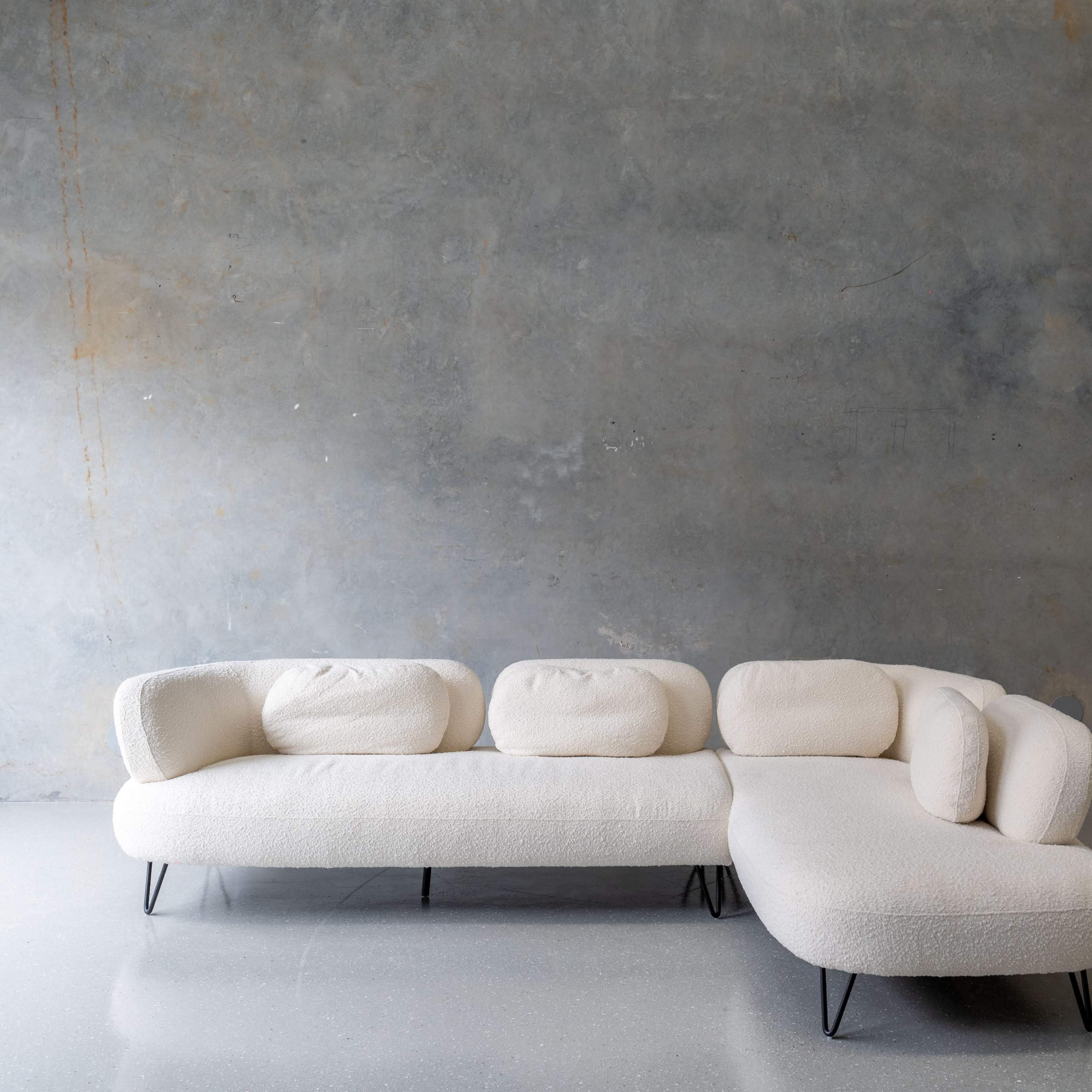 A light-colored, curved sectional sofa with rounded cushions is positioned against a textured gray wall.