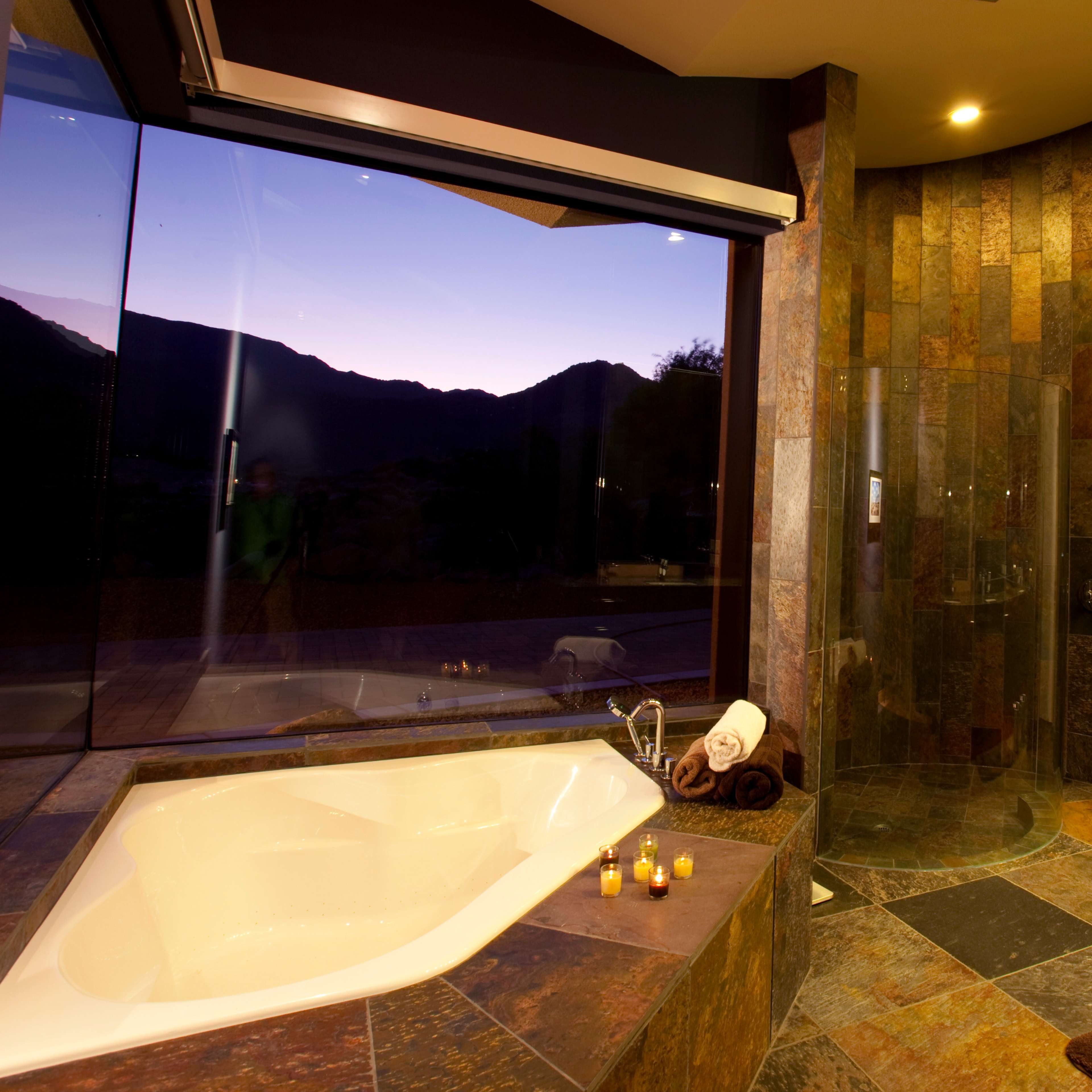 A modern bathroom with a jacuzzi tub, a glass shower, and large windows offering a view of mountains at dusk.