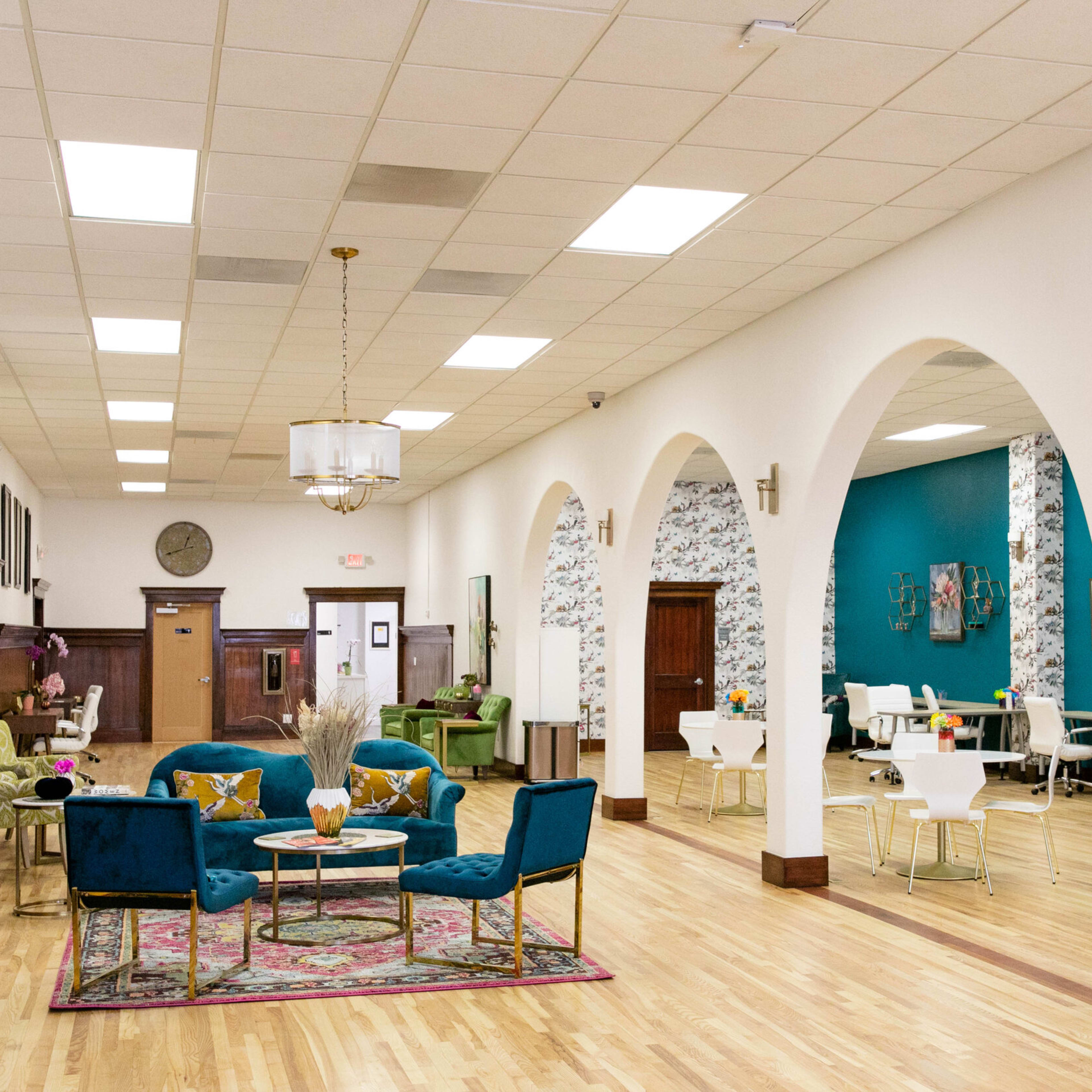A spacious, well-lit room with arched doorways, light wood flooring, and a mix of seating areas, including colorful chairs and tables arranged for dining or socializing.