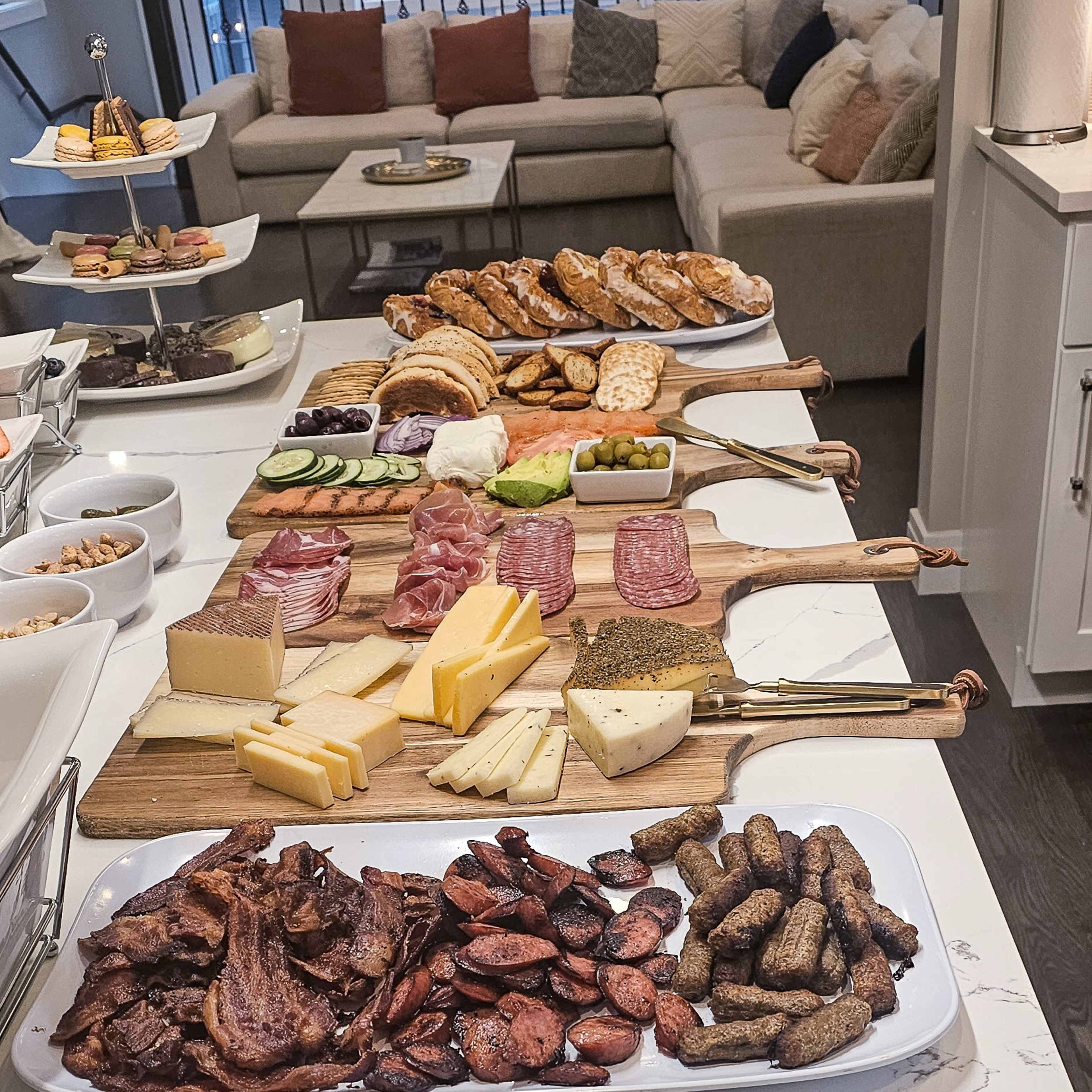 A buffet-style spread featuring assorted cheeses, cured meats, bacon, sausages, and French toast arranged on white serving platters.