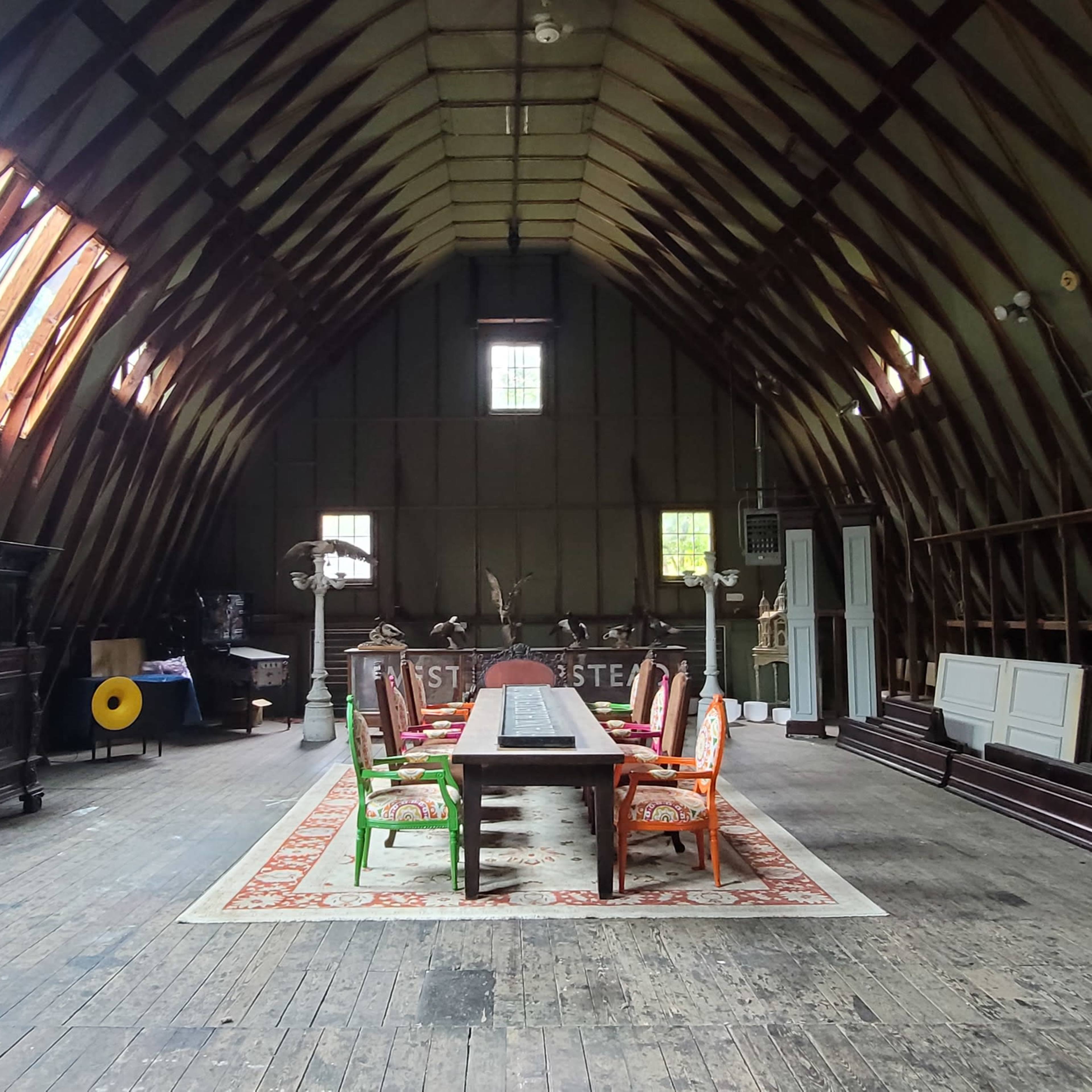 A spacious attic with wooden beams, featuring a long dining table surrounded by brightly colored chairs, an ornate circular decoration on the left, and various pieces of furniture scattered around.