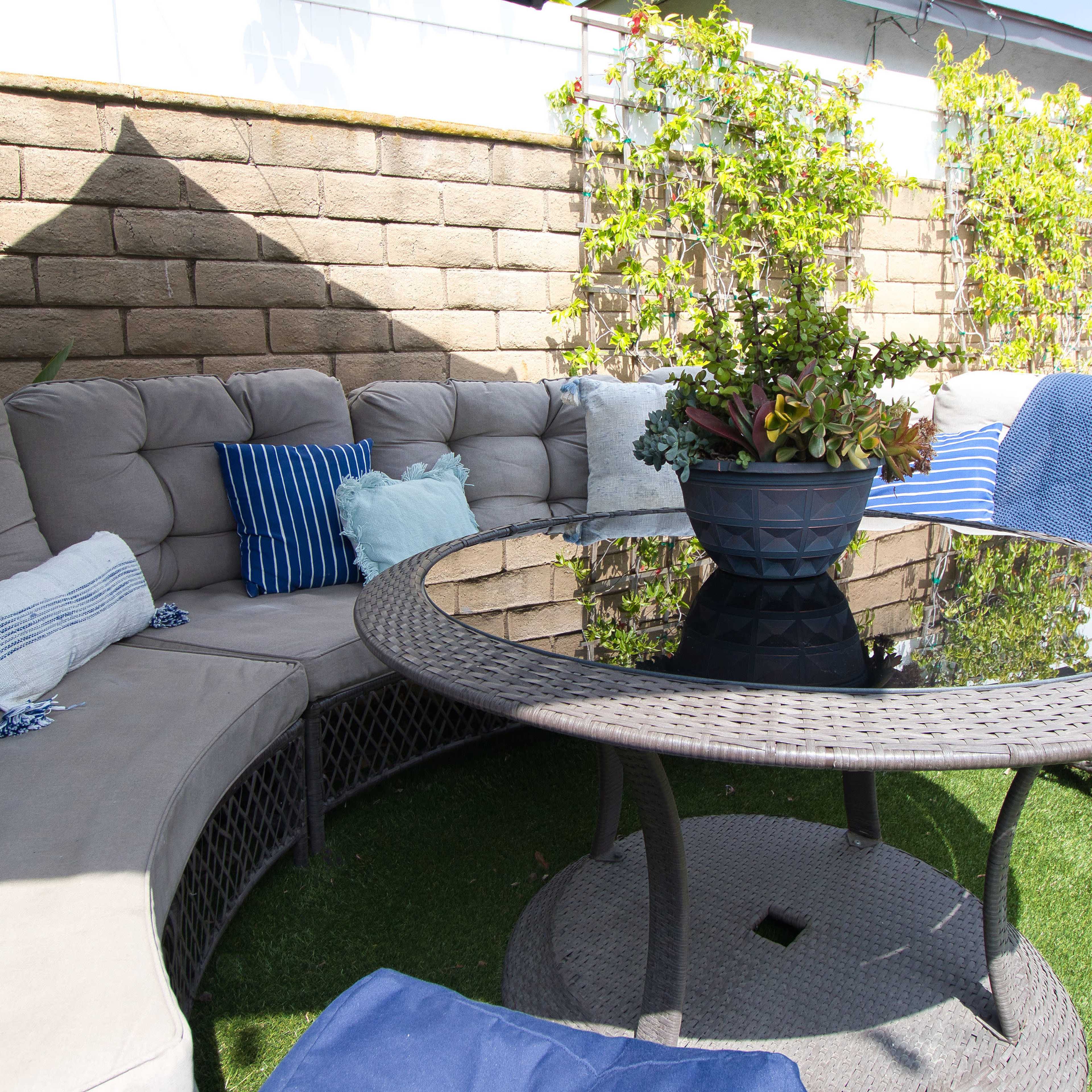 A circular seating area with a glass table is surrounded by cushions and a potted plant in a landscaped outdoor space.