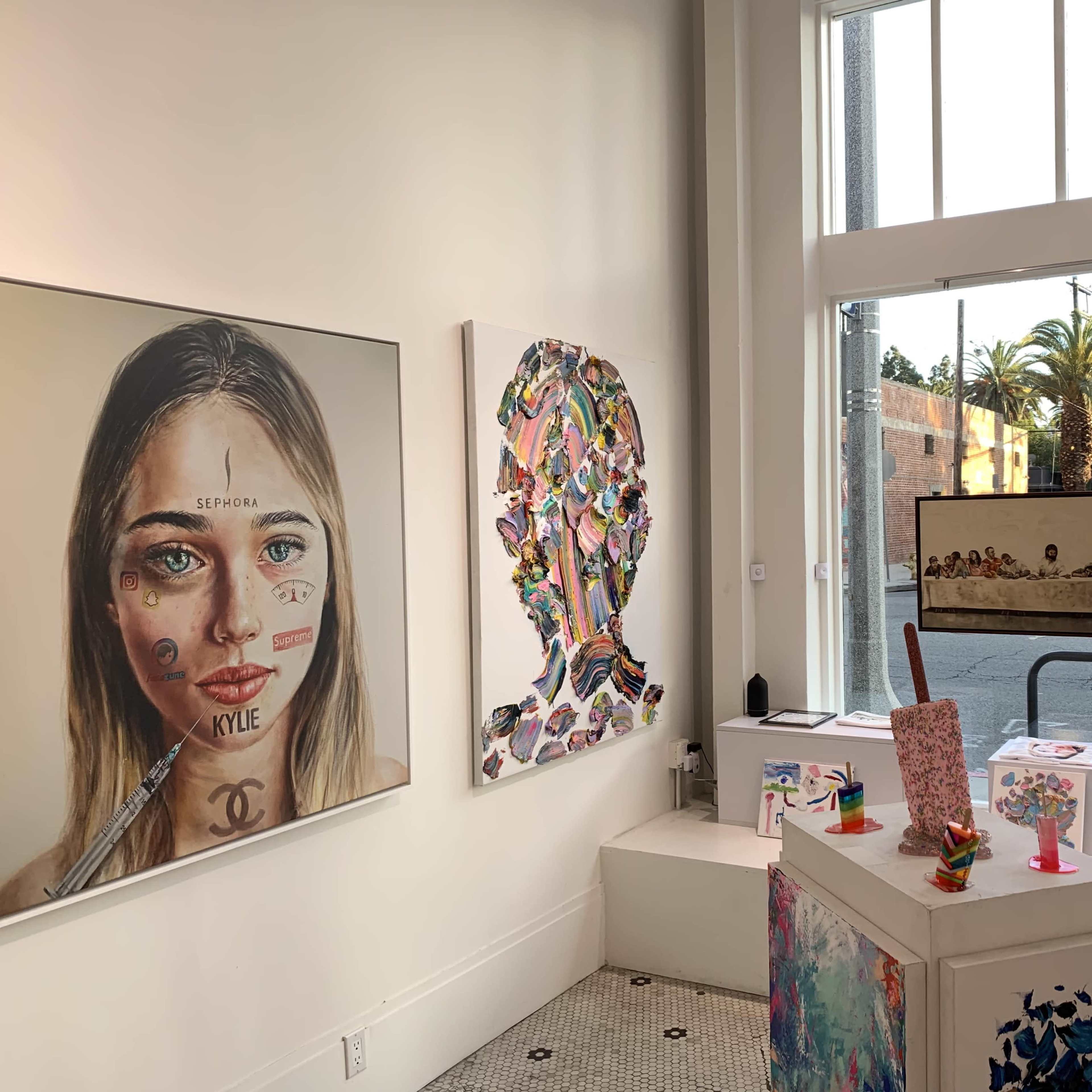 An art gallery with two prominent artworks on the walls, one of a portrait and the other abstract, alongside a display area featuring colorful sculptures and a view of palm trees outside the window.