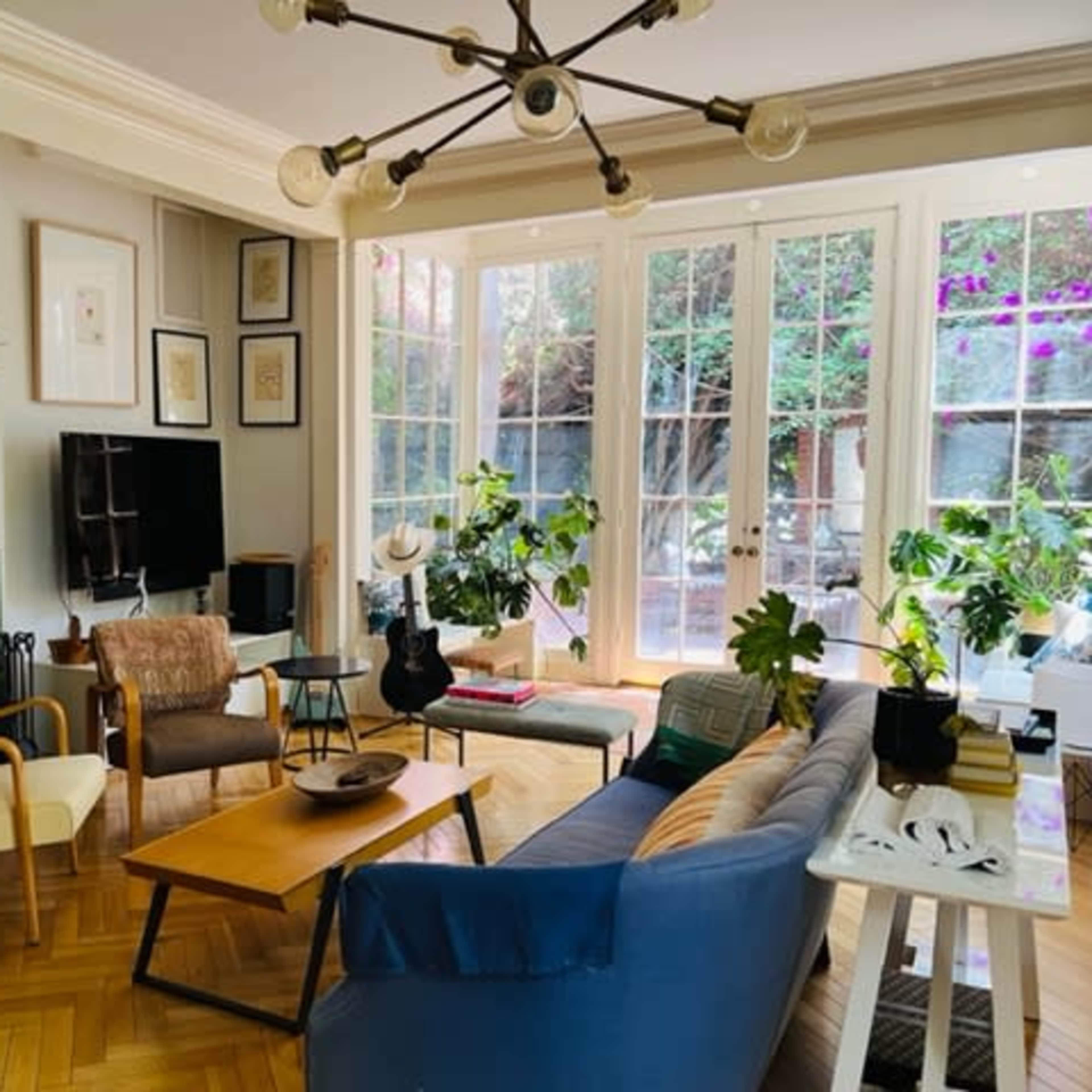 The living room features a mix of modern and vintage furniture, large glass doors leading to a garden, and several indoor plants.
