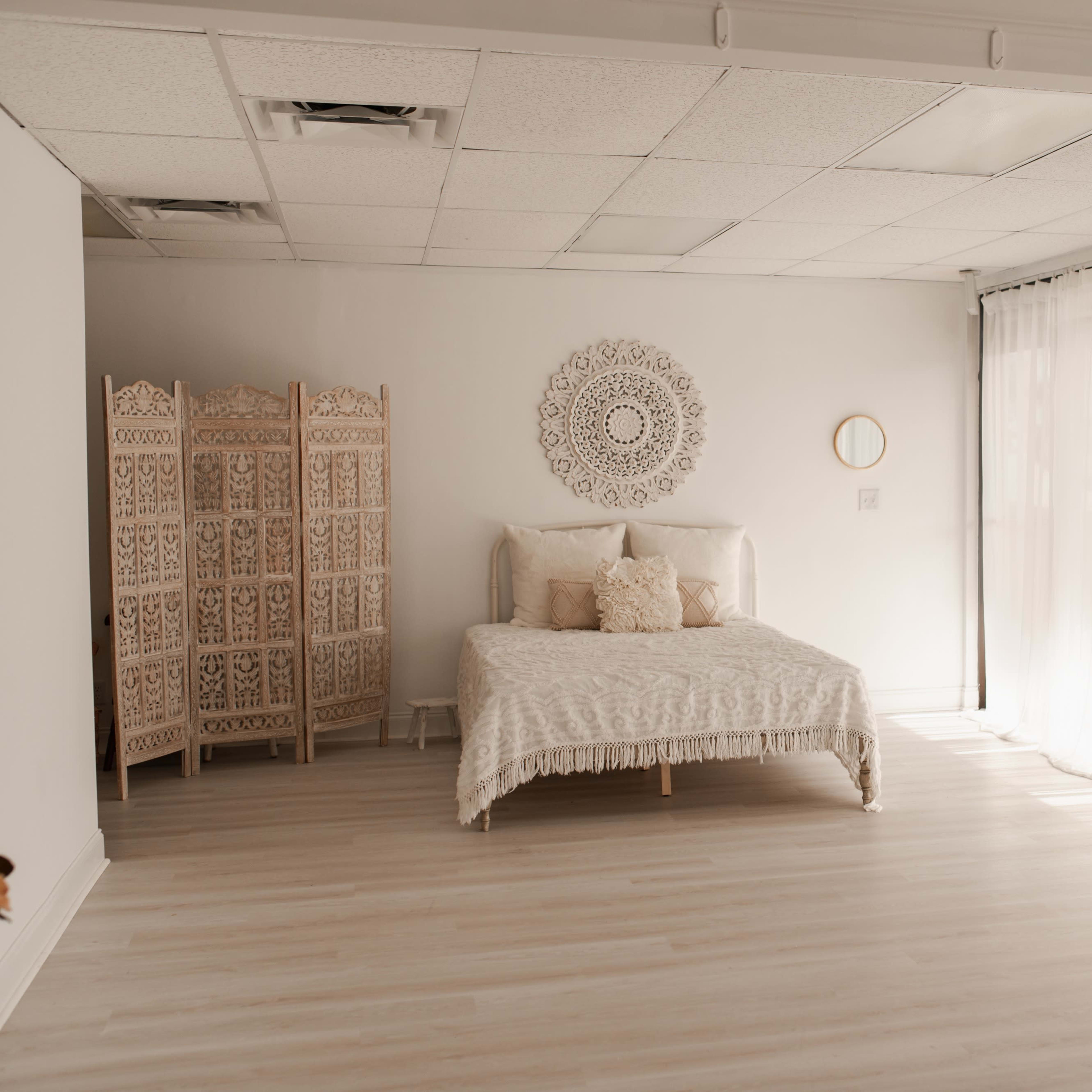 A minimalist bedroom featuring a bed with light bedding, a decorative wall piece, and large windows covered with sheer curtains allowing natural light to fill the space.