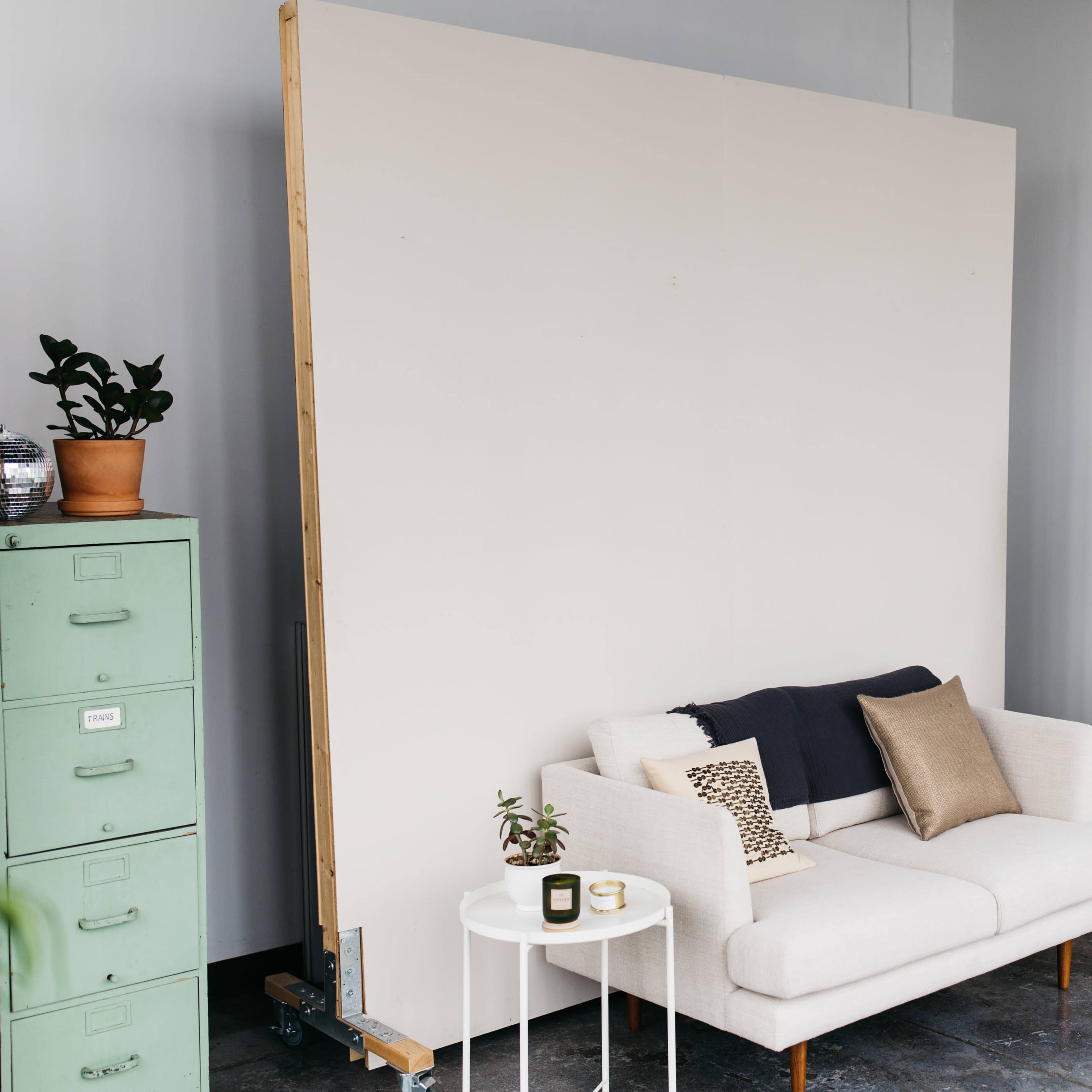 A minimalist interior with a white sofa, a side table with a candle, a large blank wall, a full-length mirror, and potted plants.