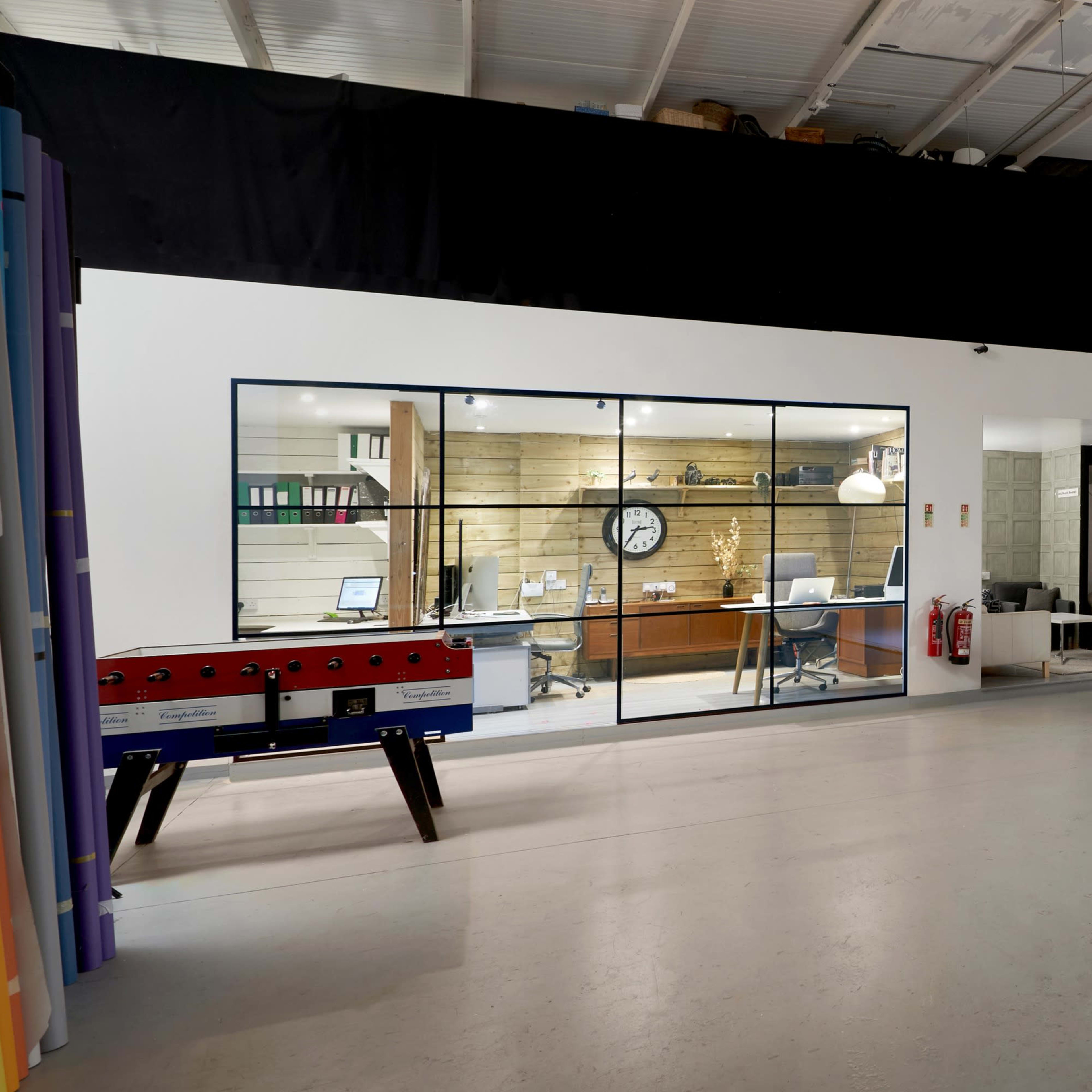A spacious, modern office area with a glass-walled workspace on one side and an empty photography studio setup on the other.