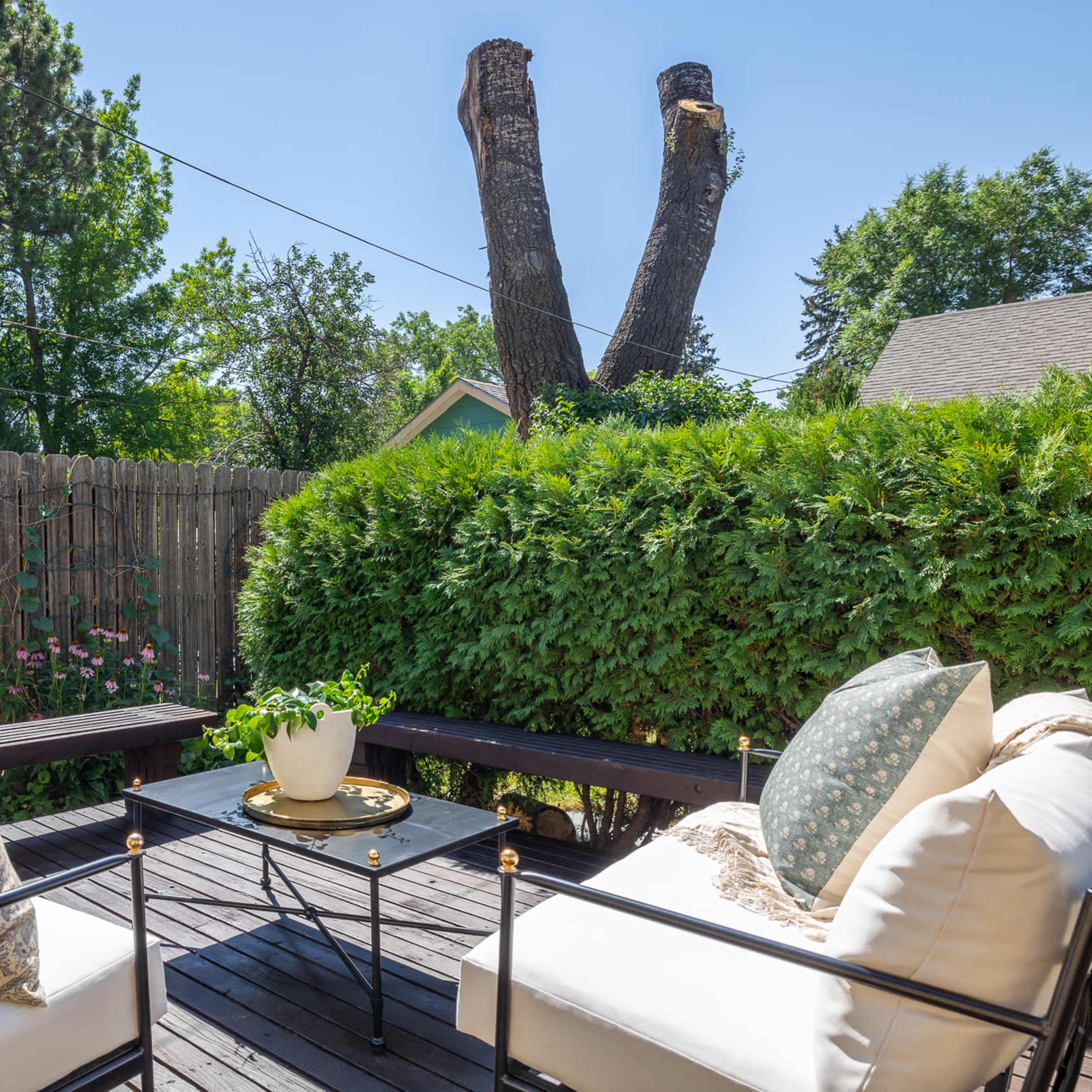 A seating area with white cushions and a small table is set on a wooden deck surrounded by greenery and a wooden fence, with two tall, cut trees in the background.