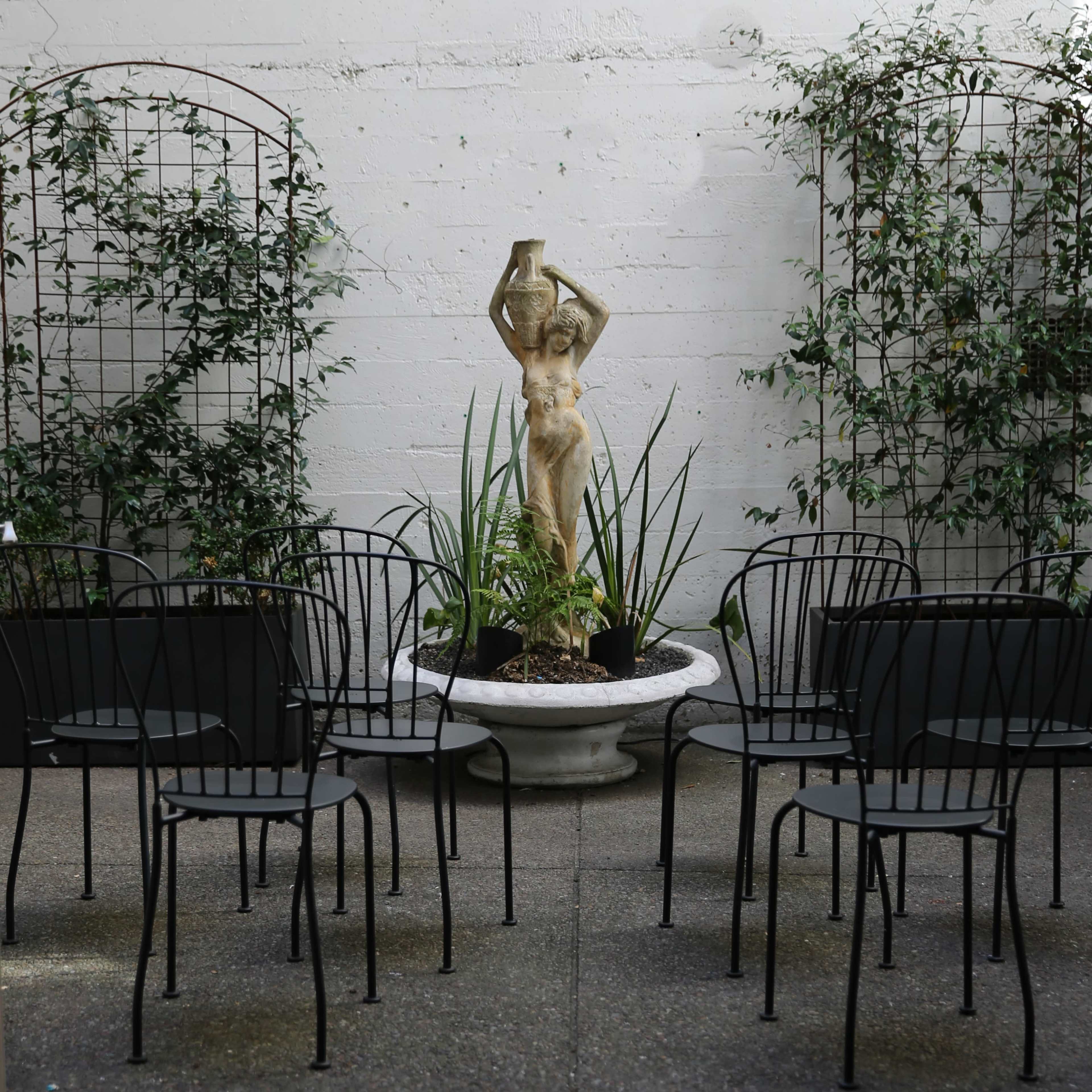 A stone statue of a woman holding a vessel stands in a planter surrounded by black chairs and greenery against a white wall.