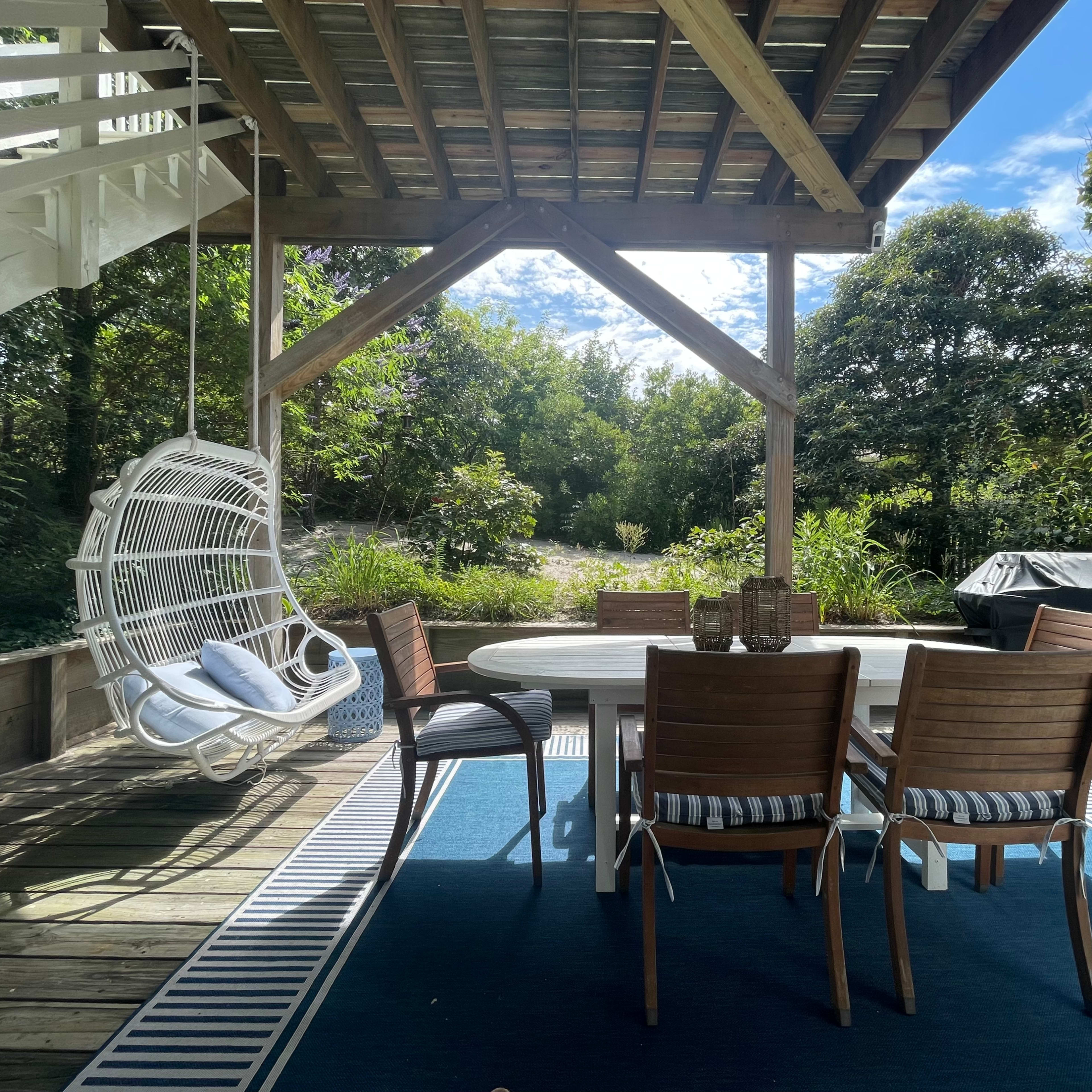 An outdoor dining area features a round table surrounded by chairs, with a hanging chair nearby, all situated under a wooden shelter amidst greenery.