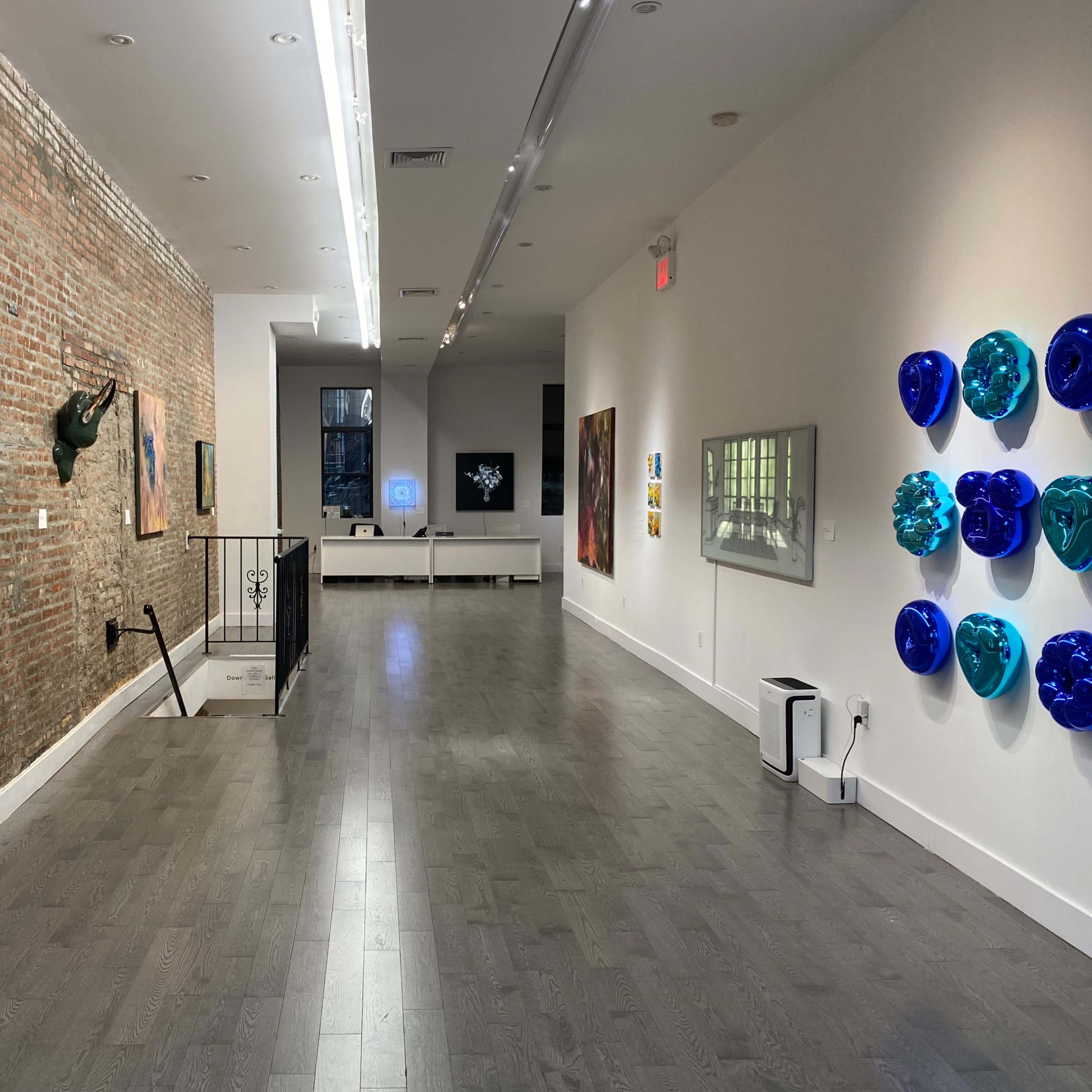 A contemporary art gallery with brick walls featuring various artworks, a staircase, and an area for displaying multimedia pieces.