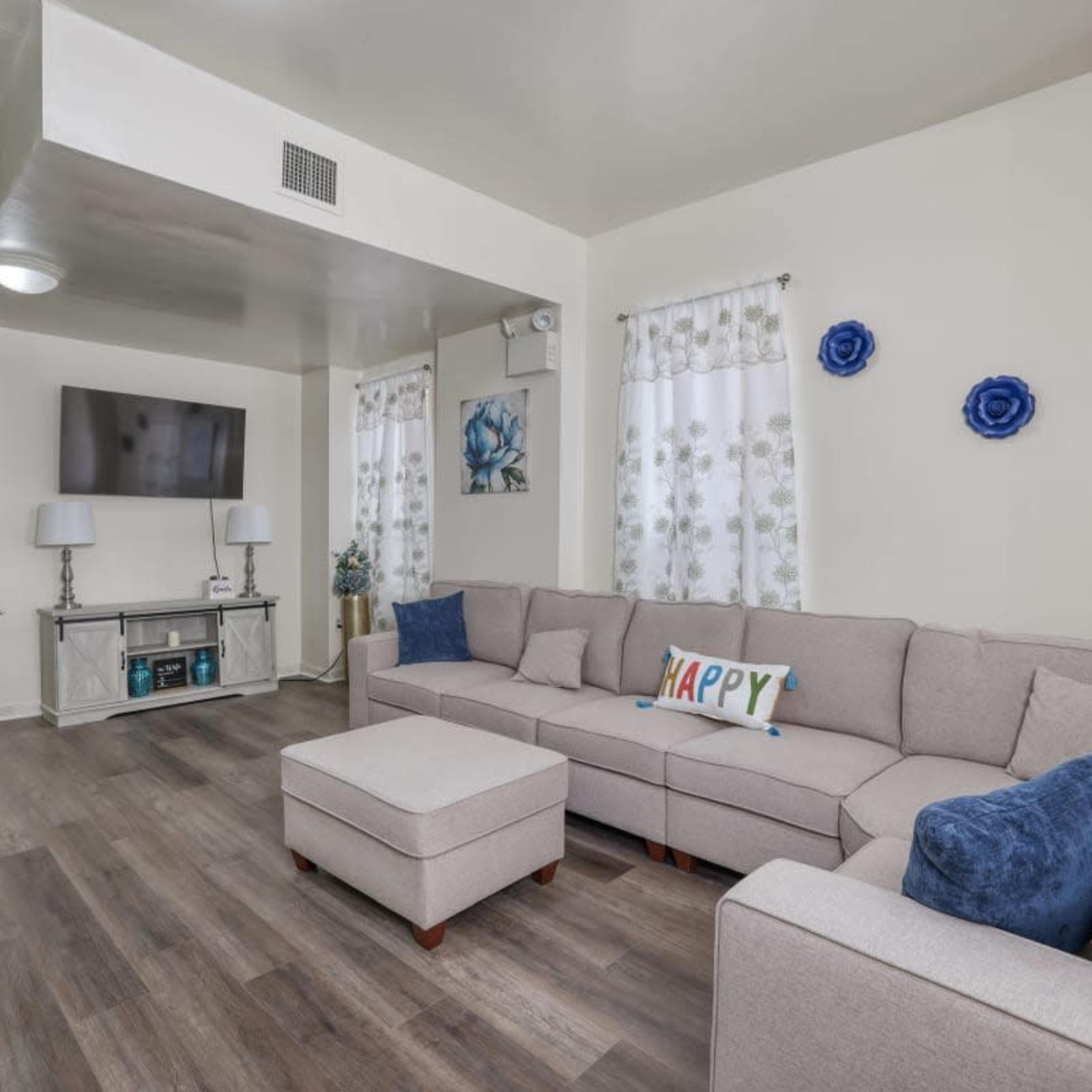 A modern living room with a gray sectional sofa, an ottoman, a television on the wall, and decorative plants.