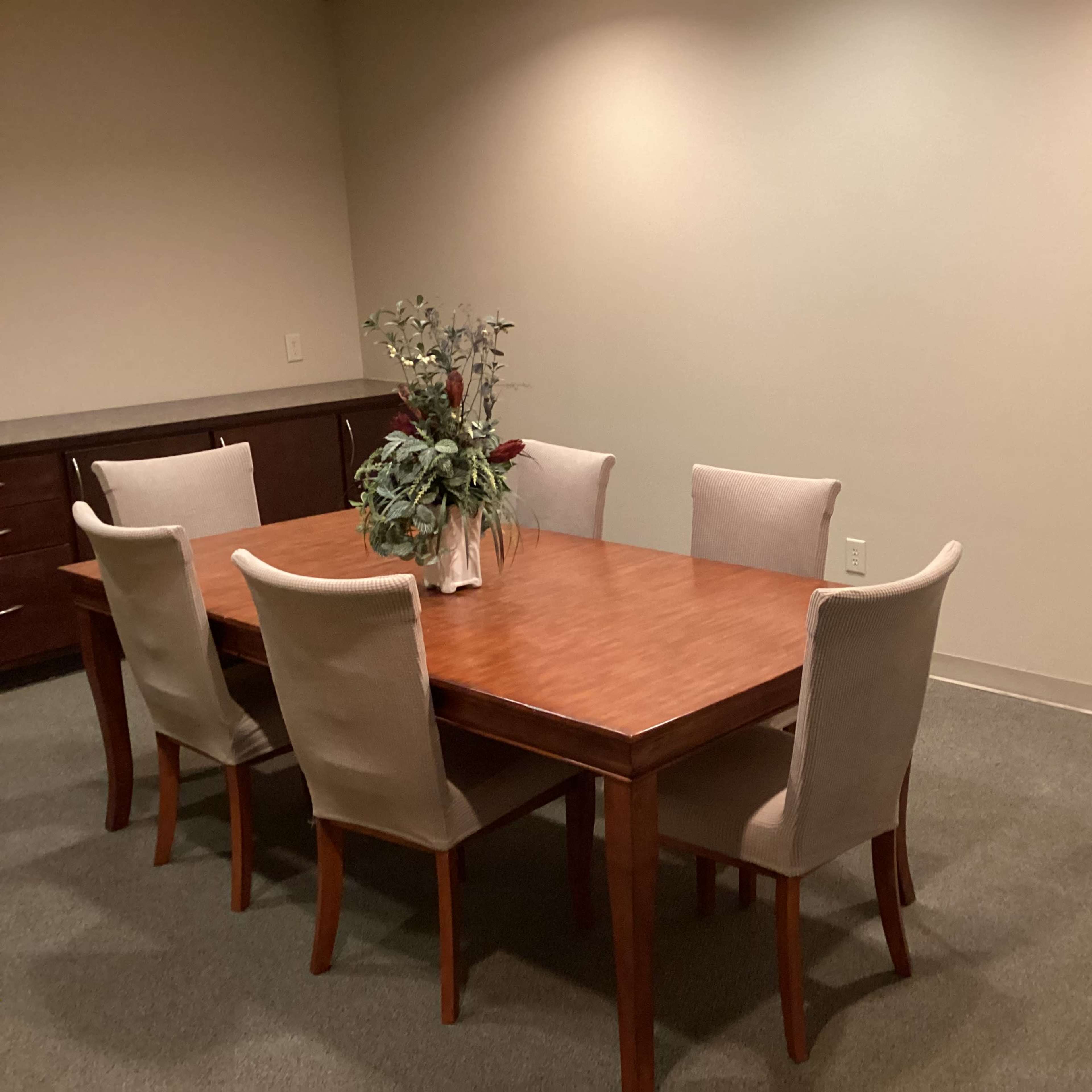 A wooden dining table surrounded by six chairs is set in a sparsely furnished room, with a potted plant at the center and a tripod in the corner.